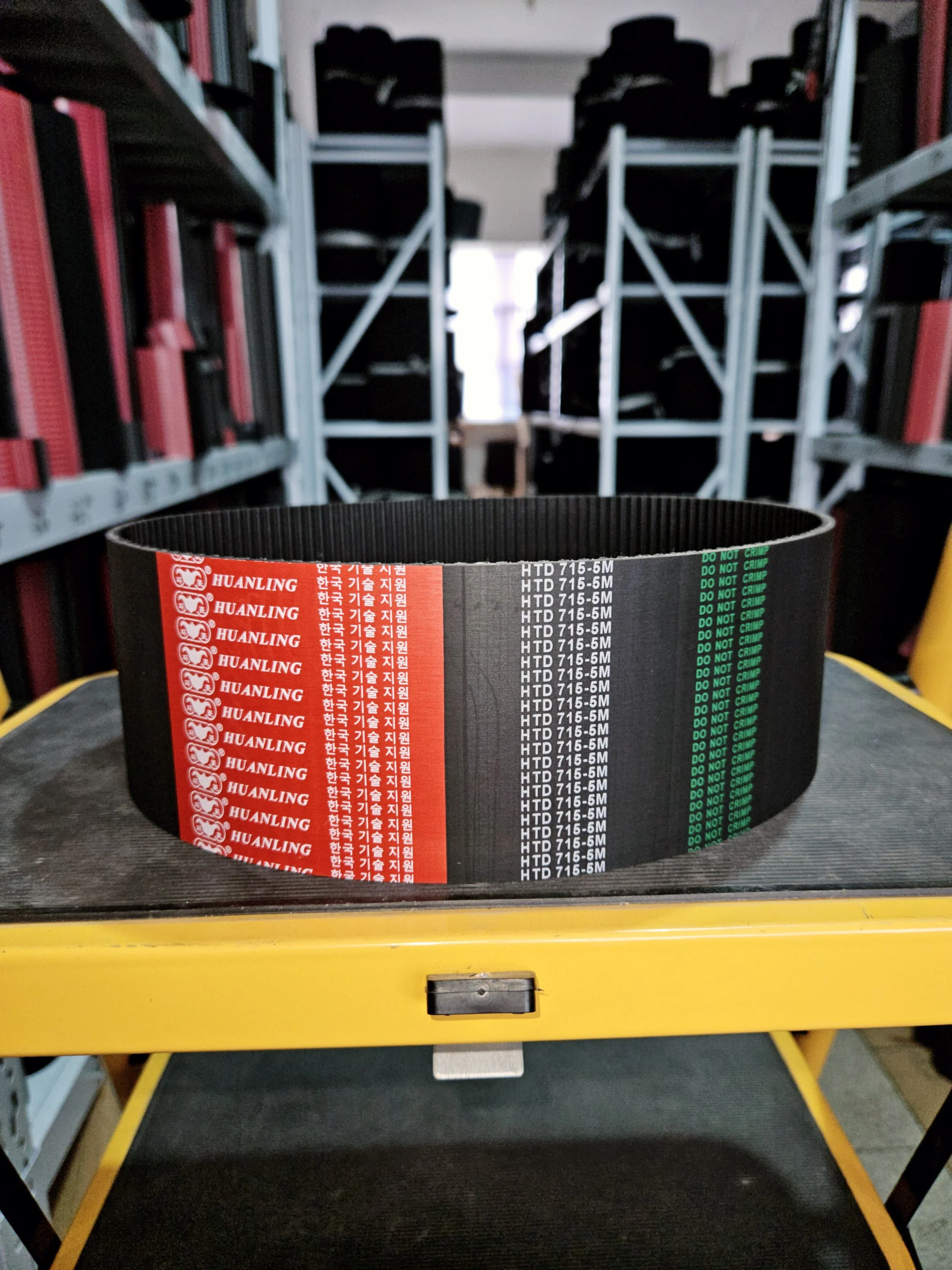 A black timing belt with red, white, black, and green labels, placed on a yellow shelf in a warehouse or workshop.