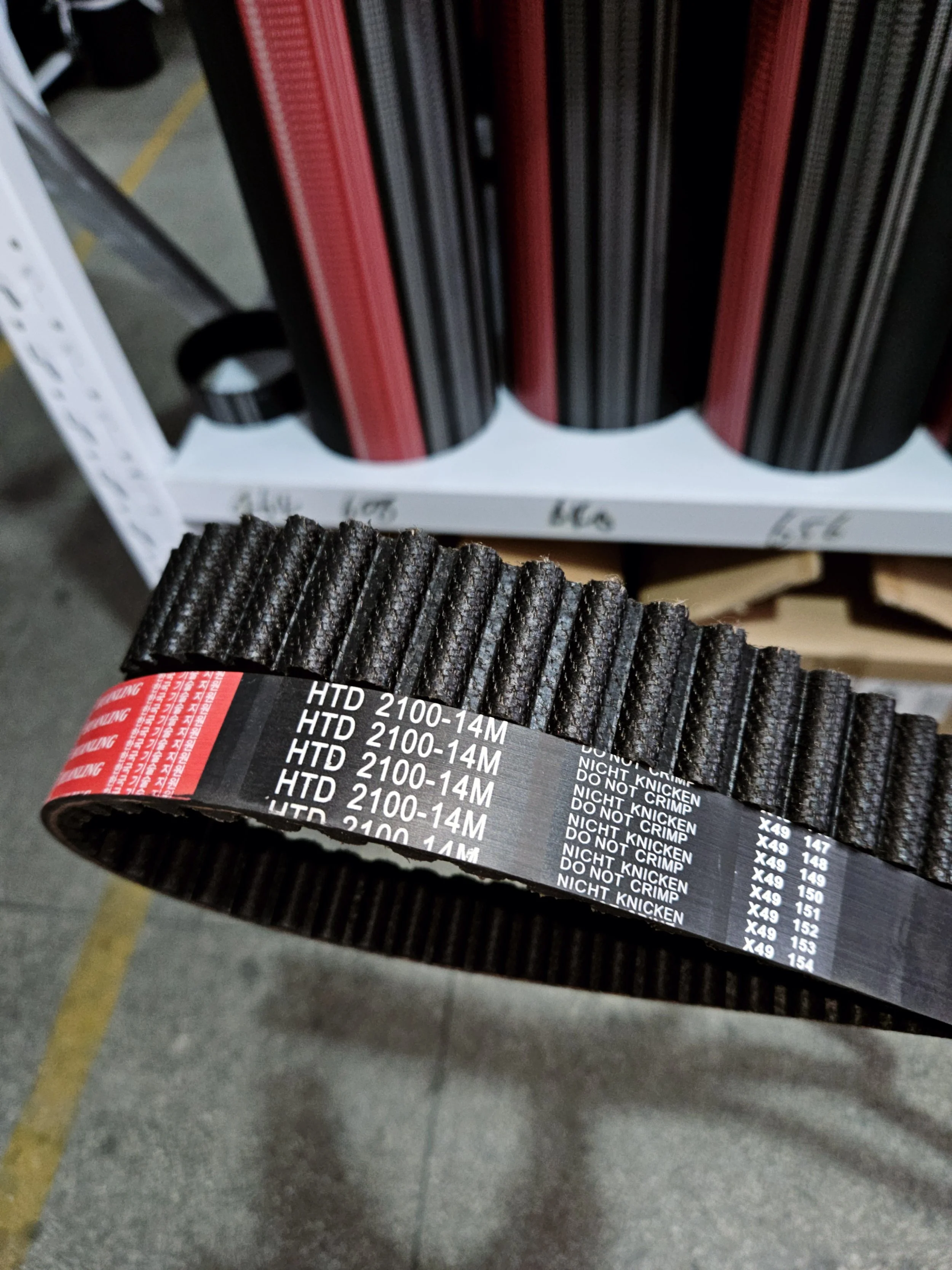 A close-up of a black rubber belt with white printed text, held in a person's hand in a hardware or store setting with shelves of similar belts in the background.