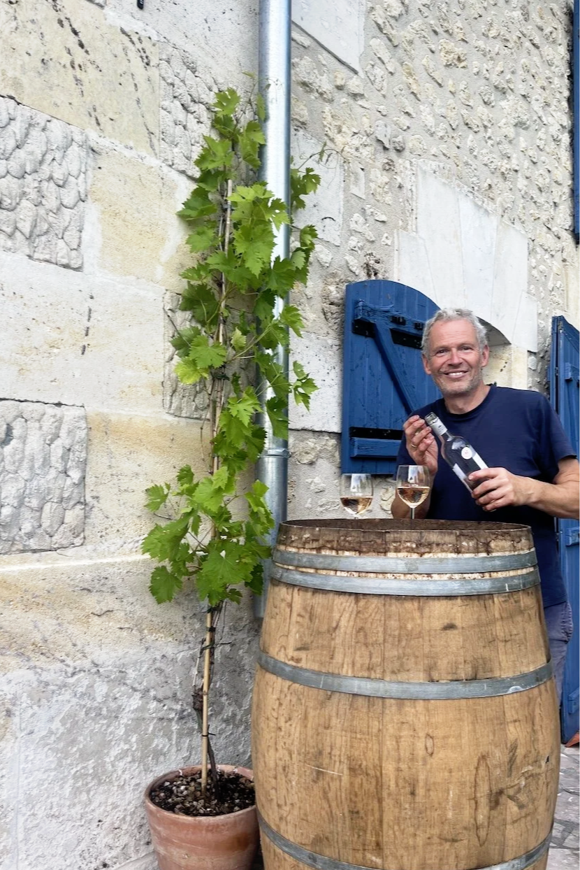 Gastgeber hinter einem Weinfass mit Weinreben, hält eine Weinflasche und zwei gefüllte Weingläser