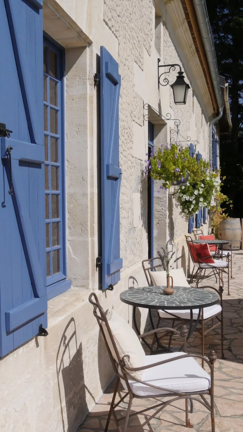 Seitenansicht eines Landhauses mit blauen Fenstern und Gästetischen mit Stühlen auf der Terrasse