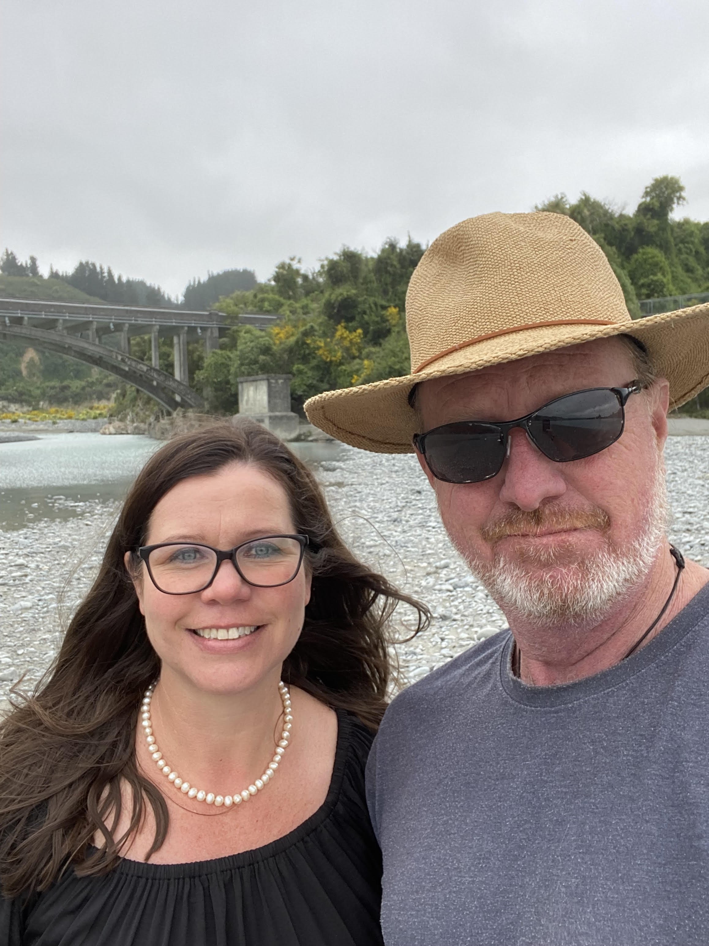 A smiling woman with long brown hair and glasses, wearing a black top and pearl necklace, standing beside a man with a beard, sunglasses, a wide-brimmed hat, and a gray t-shirt, outdoors near a river with a bridge and green trees in the background.