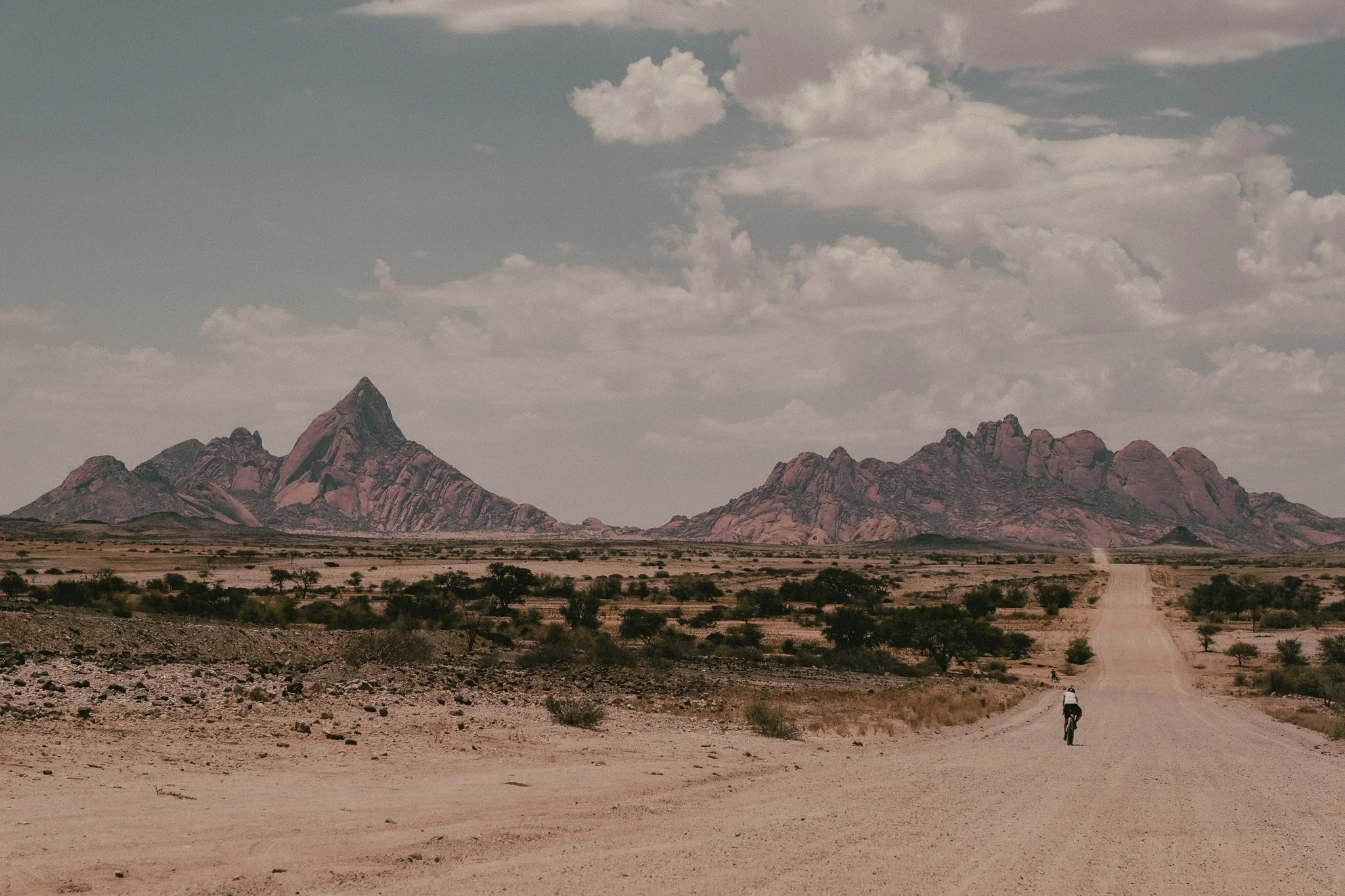 Een persoon fietst over een zandweg door een drooge, afgelegen woestijn met rotsachtige bergen op de achtergrond onder een bewolkte hemel.