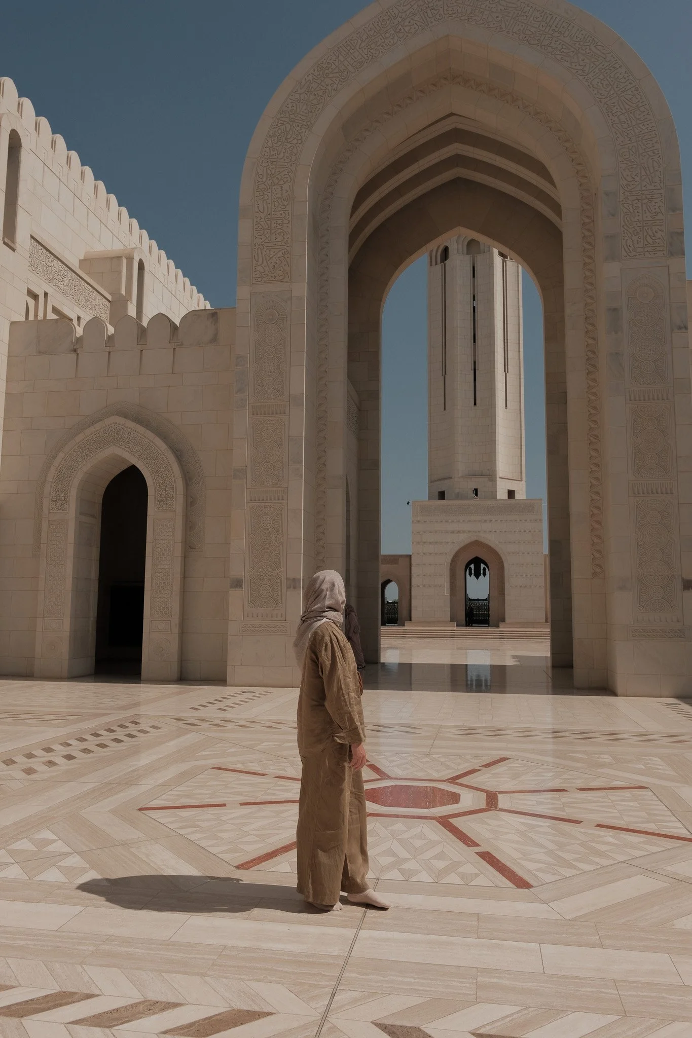 Een persoon met een hoofddoek in beige kleding staat voor een witte, marmeren moskee met grote poorten en een hoge minaret, onder een heldere blauwe hemel.