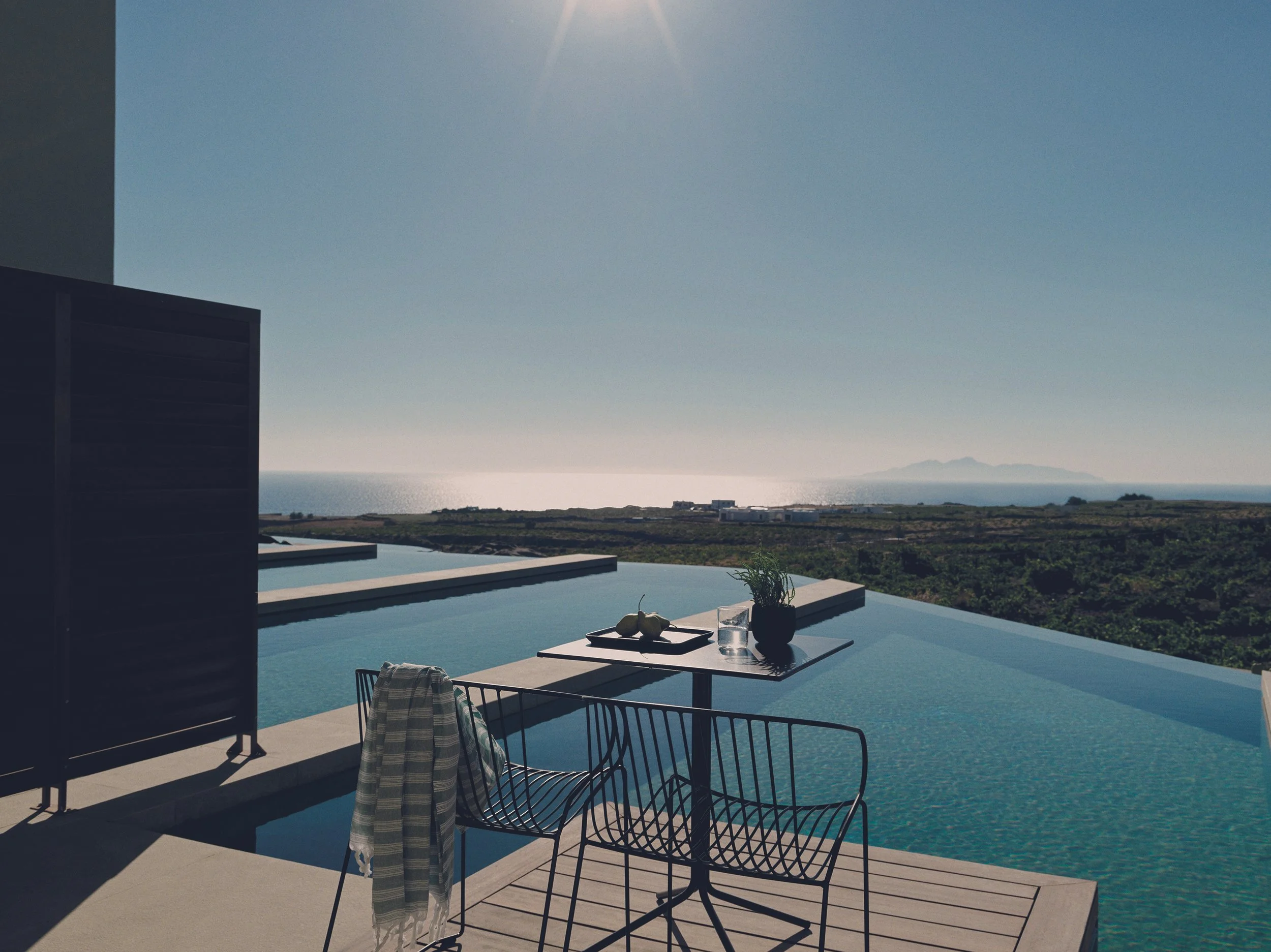 Uitzicht op de oceaan vanaf een zwembad met een tafel en twee stoelen in de tuin. Op de tafel liggen een wijnglas, een bord met groenten en een pot met planten.