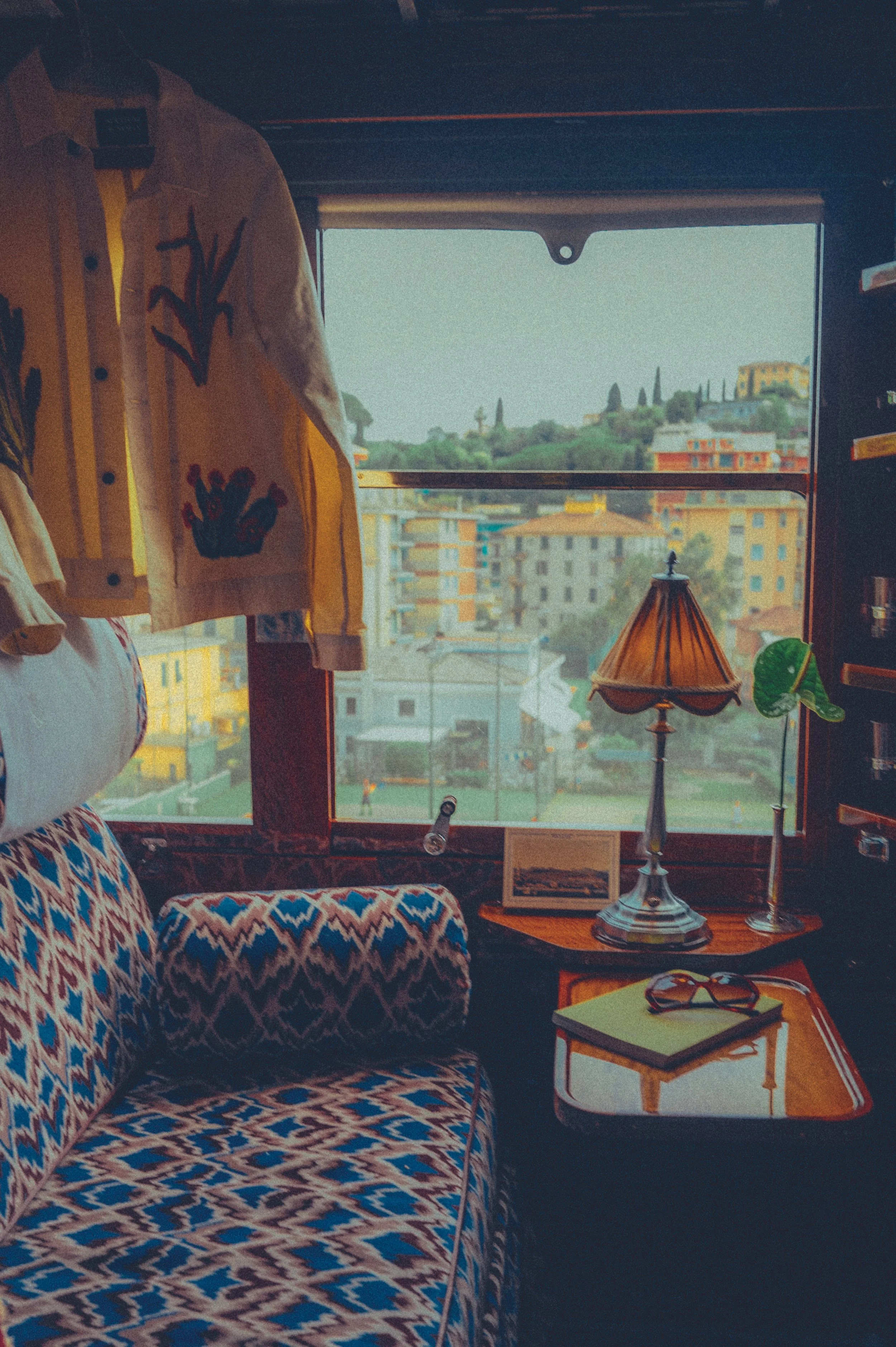 Binnenzicht van een gezellige kamer met een bank in diachroompatroon, een tafeltje met een boek en een zonnebril, een lamp en een plant, met een groot raam dat uitzicht biedt op een heuvelachtig stadslandschap met kleurrijke gebouwen en bomen.