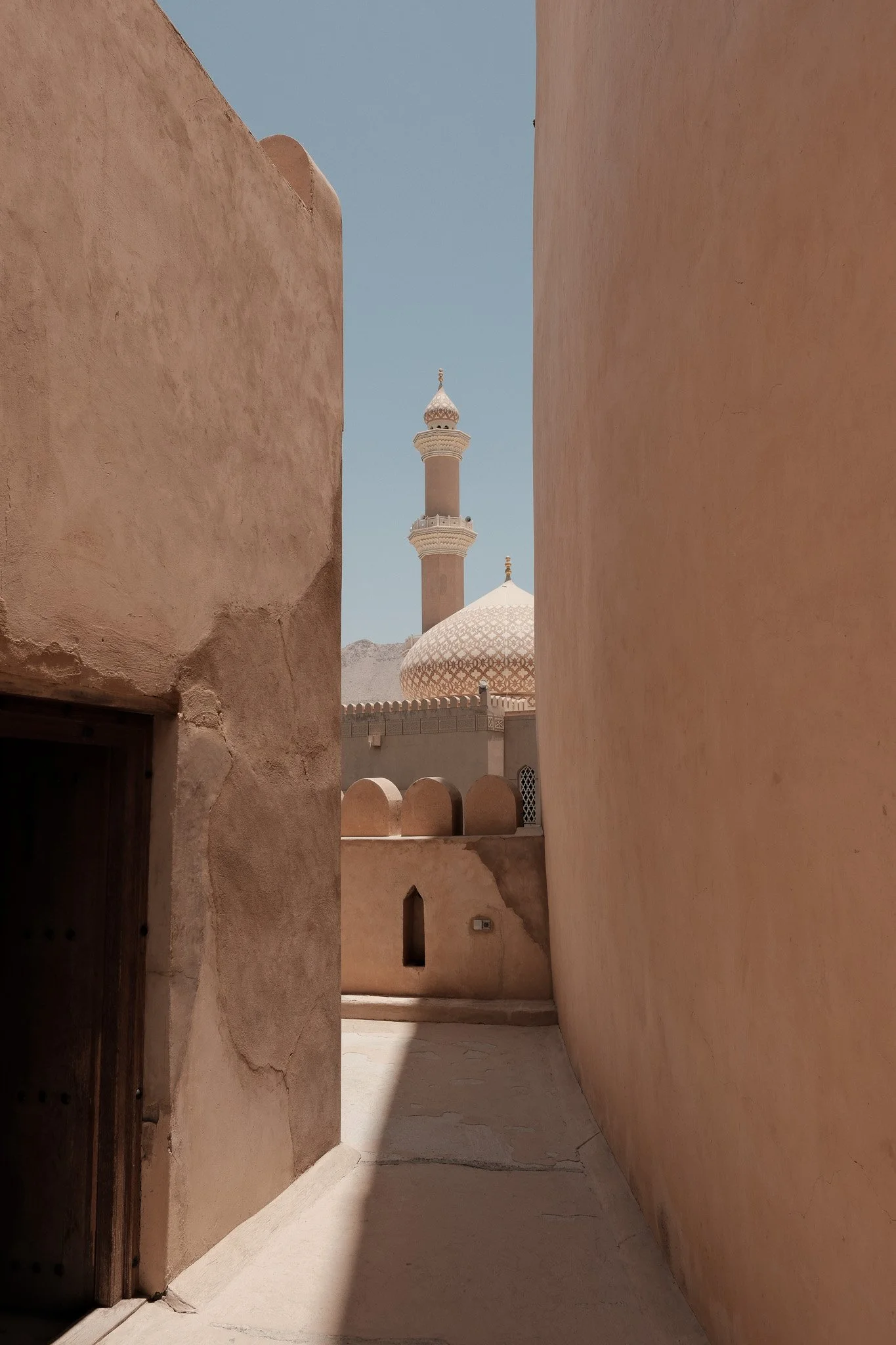 Uitzicht op een minaret en een koepel van een moskee, gezien door een smal steegje met zandkleurige muren onder een blauwe hemel.
