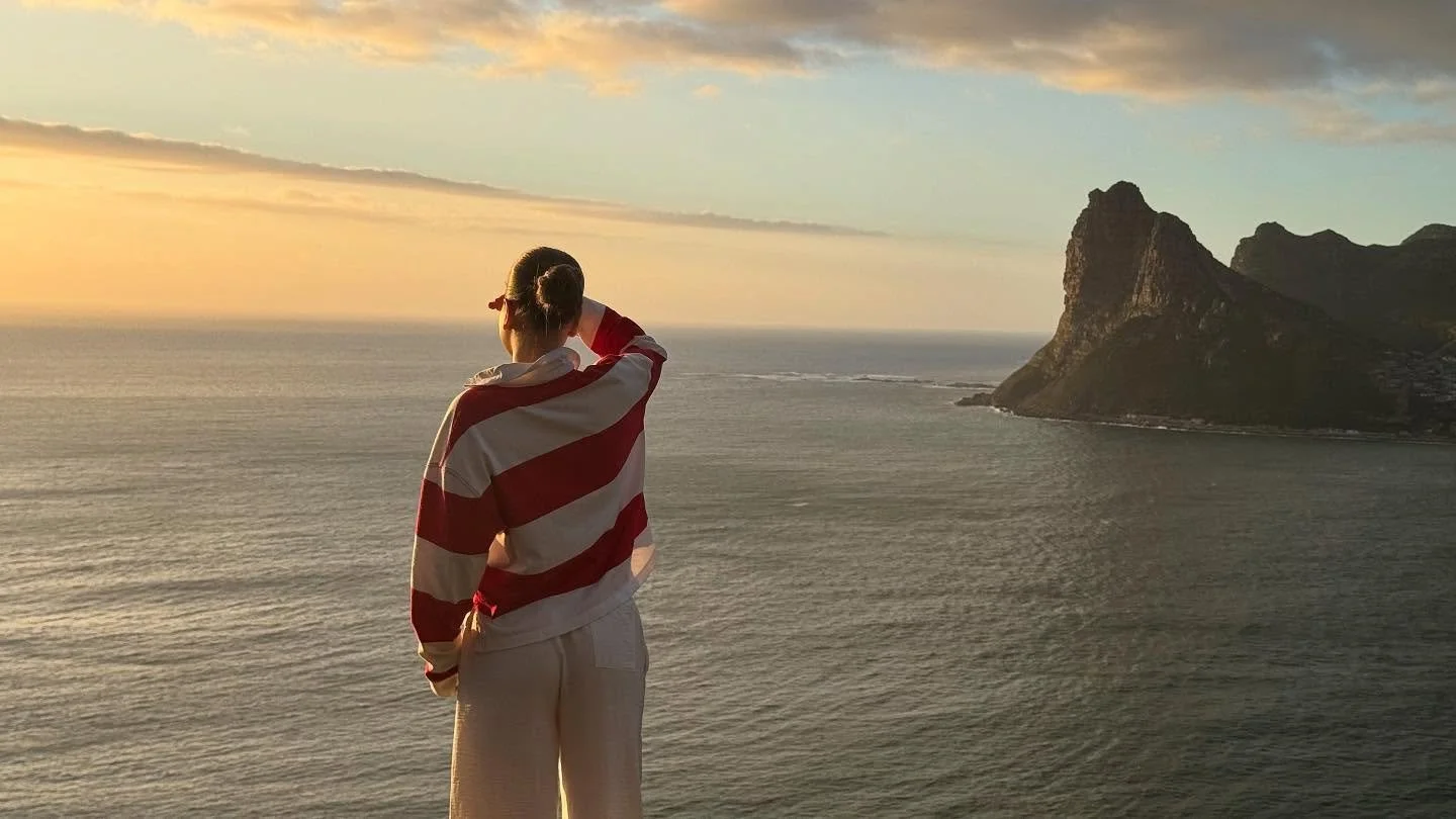 Vrouw die uitkijkt over de oceaan tijdens zonsondergang, met een roodgeblokten jas en een rotsachtig eiland op de achtergrond.