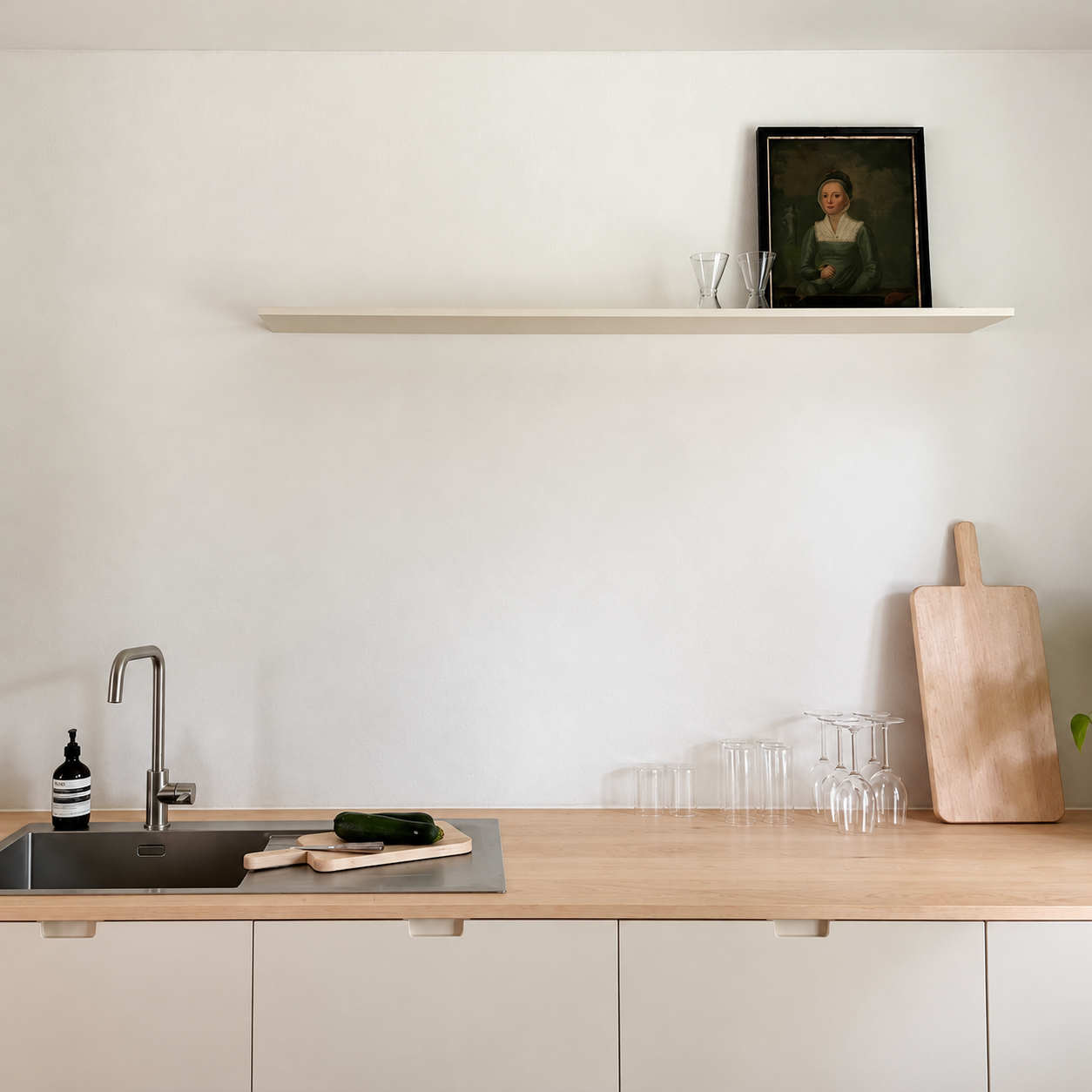 Een minimalistische keuken met een houten aanrecht, een spoelbak, een plant, een snijplank, een stuk komkommer, een fles zeep, glazen, een houten snijplank, en een muur met een zwevende plank met een schilderij en glazen.