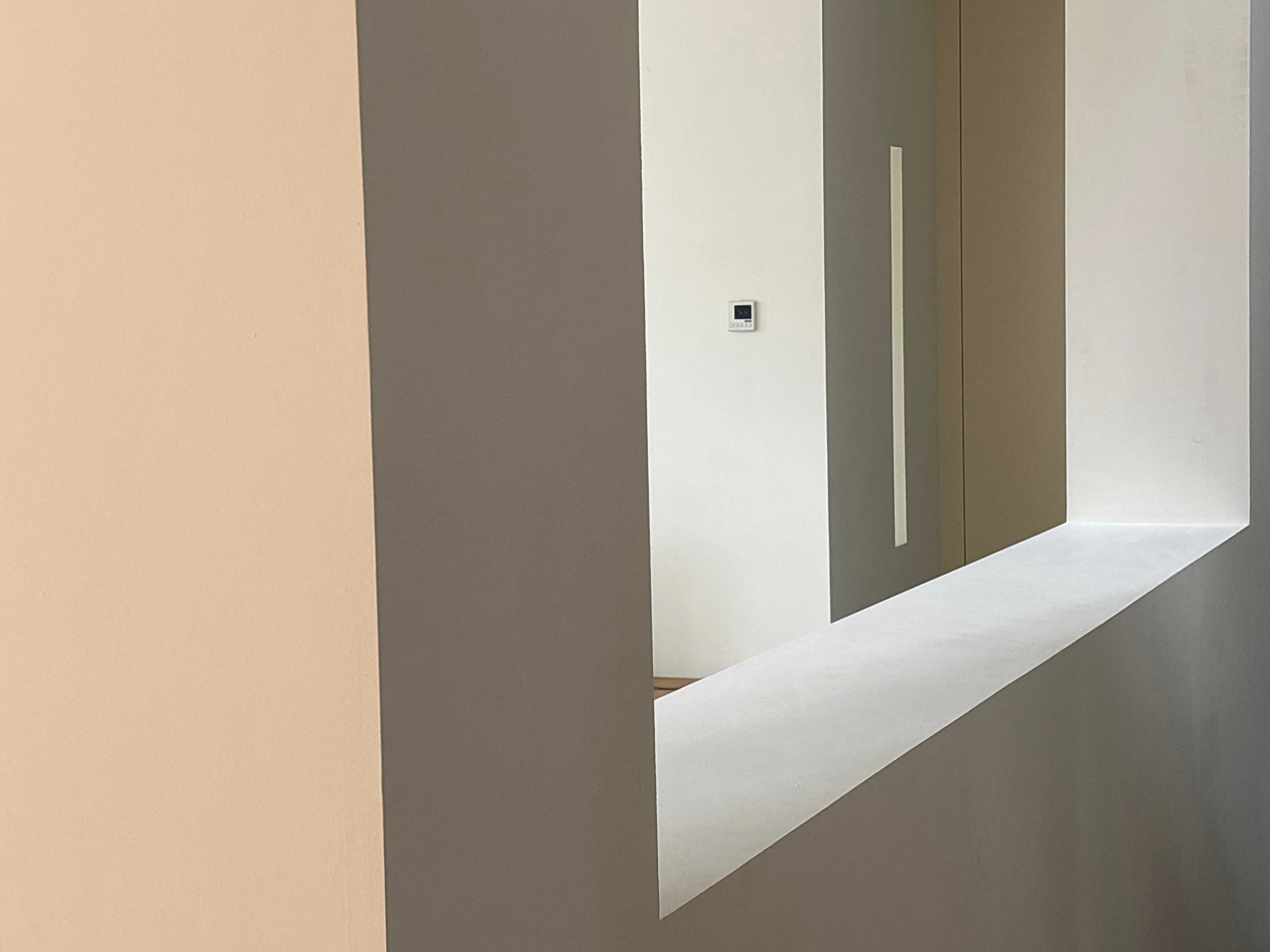 Interieur van een modern gebouw met een muur in beige, grijze, witte en houten tinten, een thermostaat op de muur en een witte rand onderaan de muur.