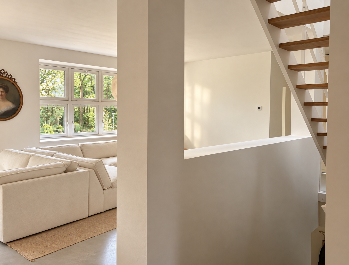 Interieur van een huis met een houten trap, witte muren en een grote raam met uitzicht op bomen. Een beige bank staat naast het raam en er hangt een portret aan de muur.