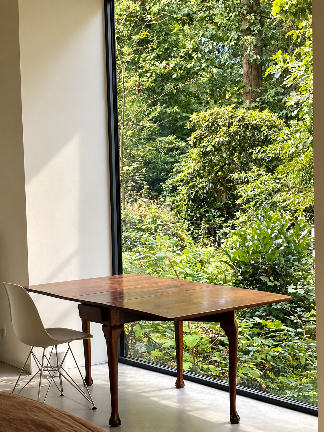 Interieur van een kamer met een houten tafel en een wit stoel naast een groot raam met uitzicht op groene bomen en planten buiten.