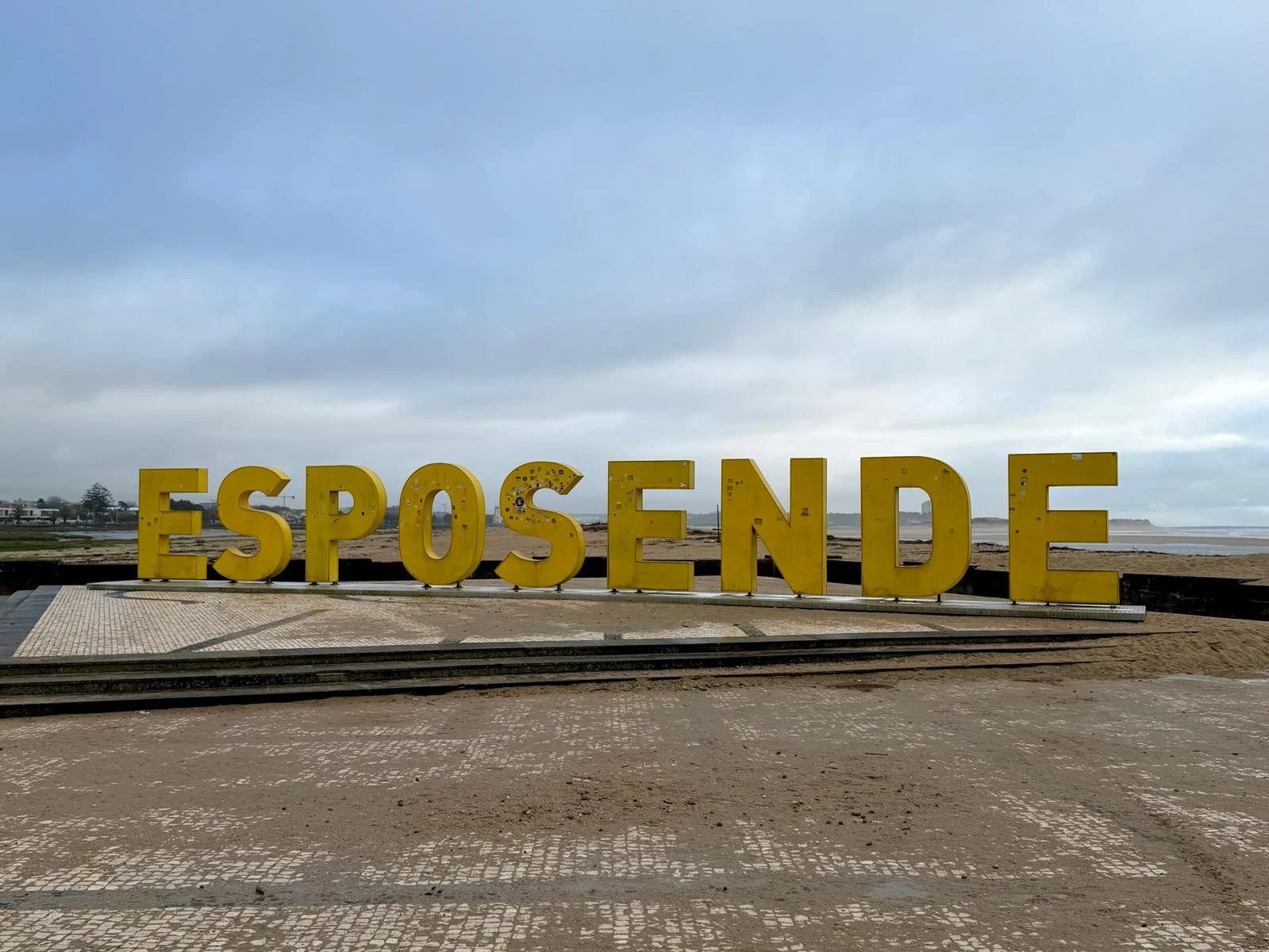 Ein großes gelbes Schild mit der Aufschrift „ESPOINDE“ an einem Strand, im Hintergrund ein bewölkter Himmel.