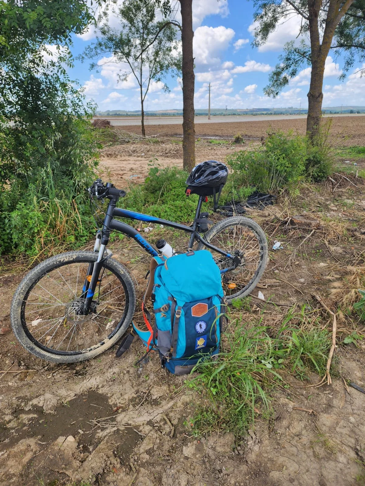 A blue mountain bike with a black helmet hanging on the seat, parked on a dirt patch beside a blue backpack with patches, under trees near a field and open sky.