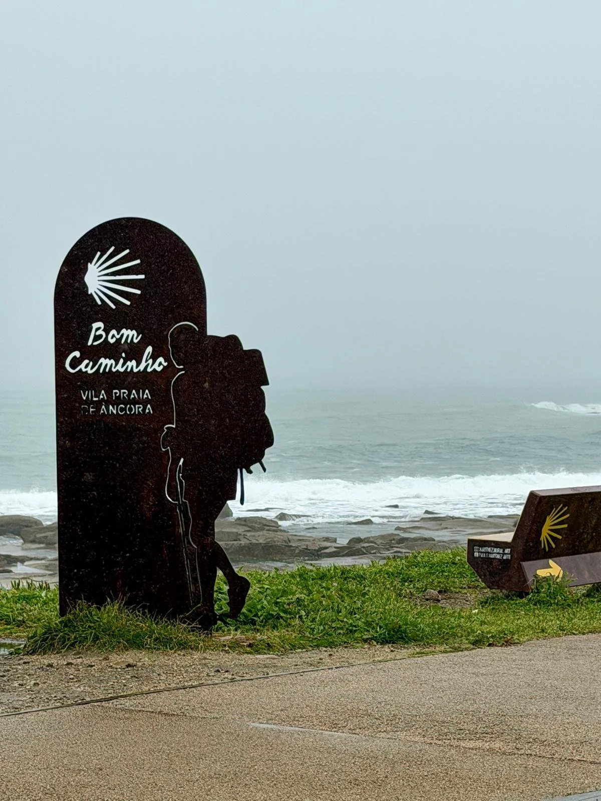 Ein Schild mit der Silhouette einer Person mit Rucksack und der Aufschrift „Bom Caminho Vila Praia de Âncora“ in der Nähe eines Strandes mit Felsen, Meer und Nebel sowie ein weiteres, kleineres Schild oder Denkmal mit einem gelben Muschelsymbol auf einer Bank.