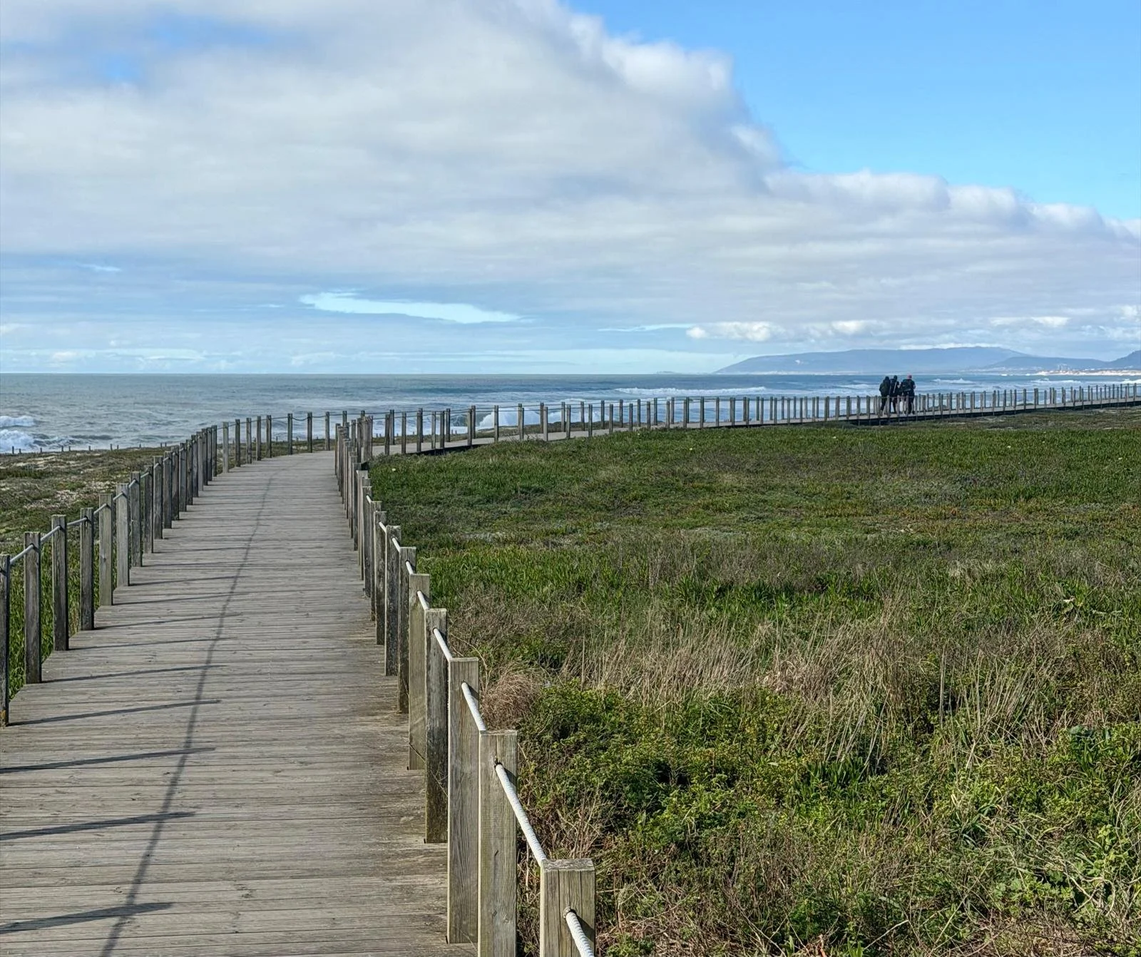 A wooden pathway with a railing leads to the beach, where a group of people walks along the shore. The sky is partly cloudy, with patches of blue, and the water is calm with gentle waves.