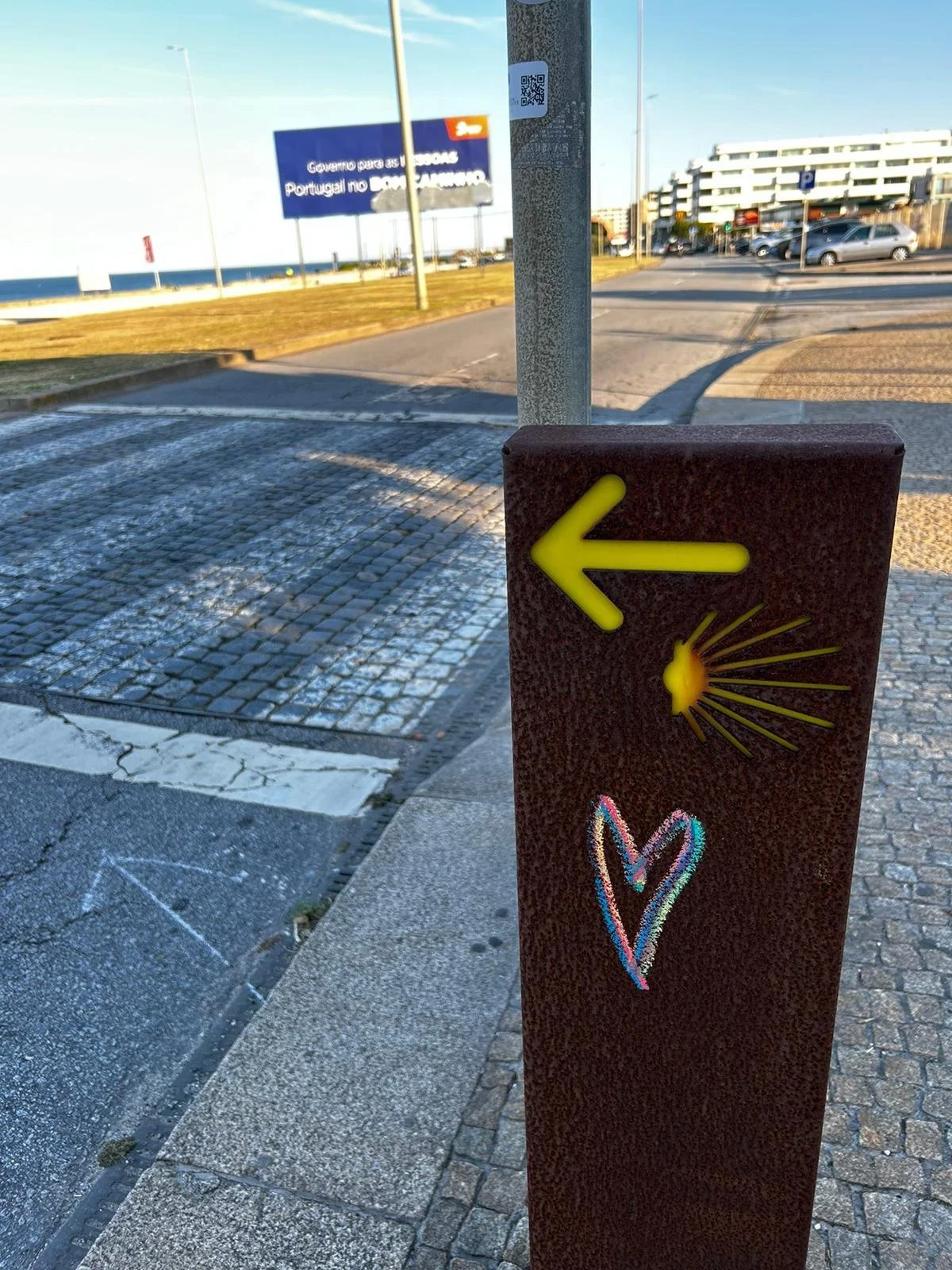 A metal post with a yellow arrow pointing left, a yellow sunburst design, and a colorful chalk heart drawing. In the background, there is a street, a parking lot with cars, and a large white building under a clear blue sky.