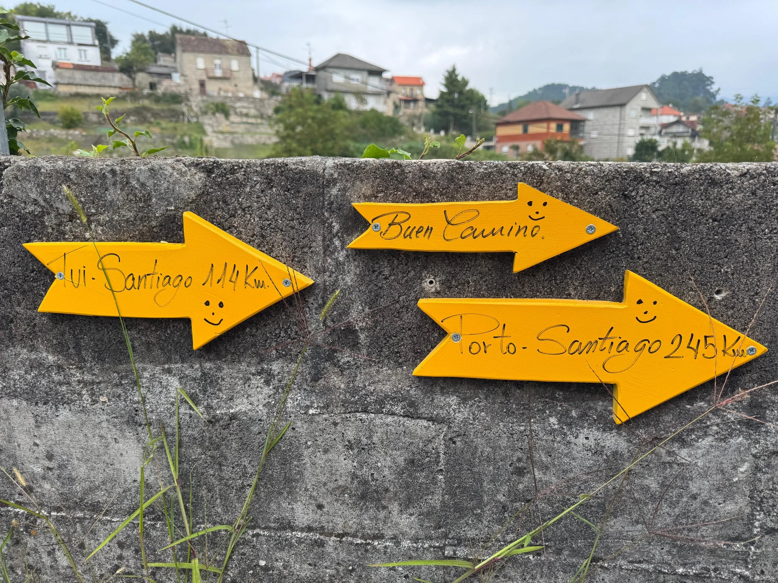Yellow directional signs on a stone wall pointing to different locations with distances. The signs say 'Tui-Santiago 114 km,' 'Buen Camino,' and 'Oporto-Santiago 245 km,' each with a smiley face drawn on them.