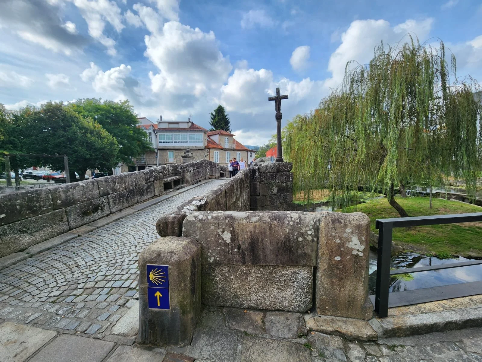 A stone bridge with cobblestone pavement and a cross on a pole, leading to a village with houses, trees, and partly cloudy sky.