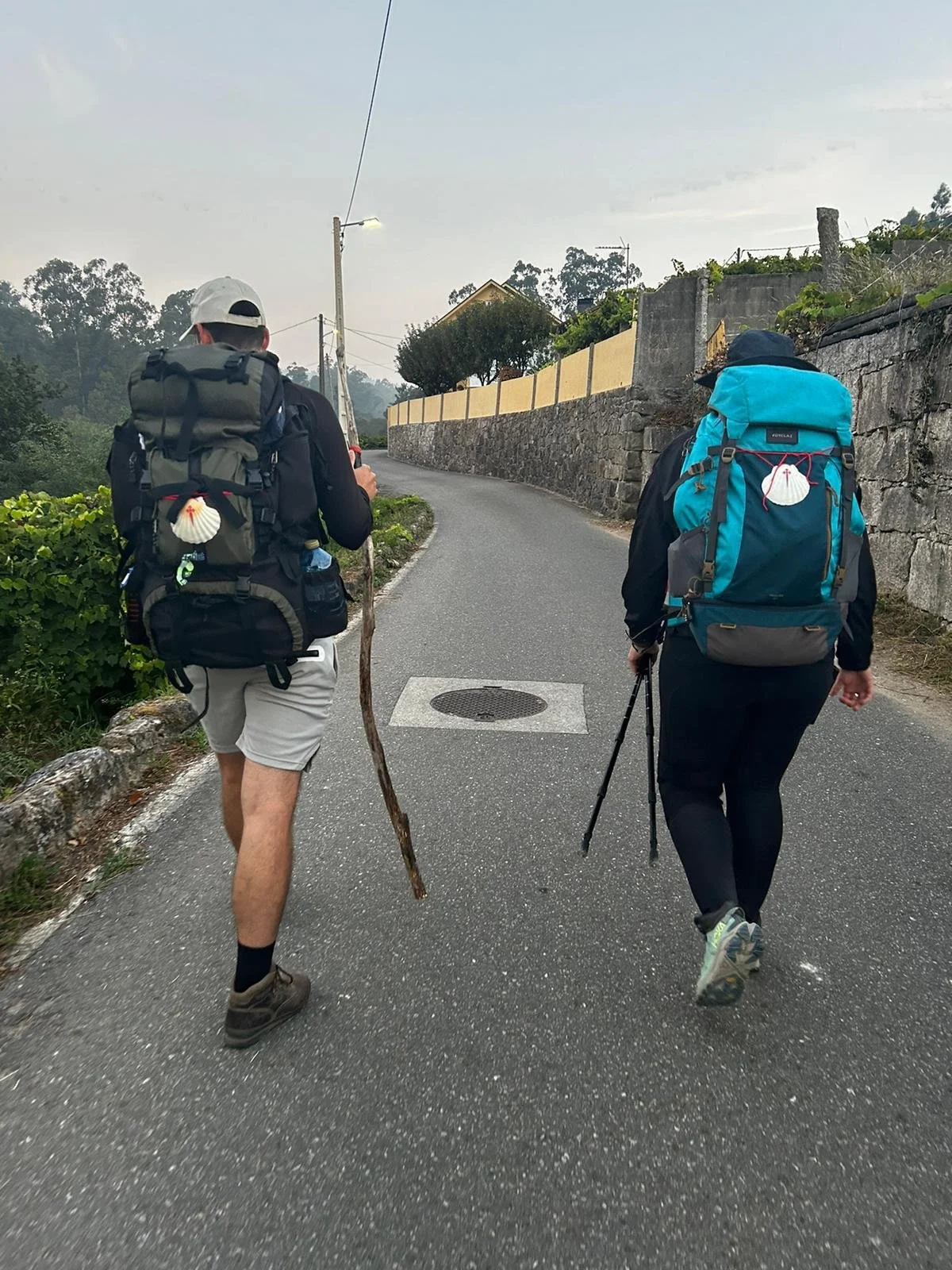 Two hikers walking on a paved road with backpacks, one holding a walking stick, in a rural area with a stone wall and trees in the background.