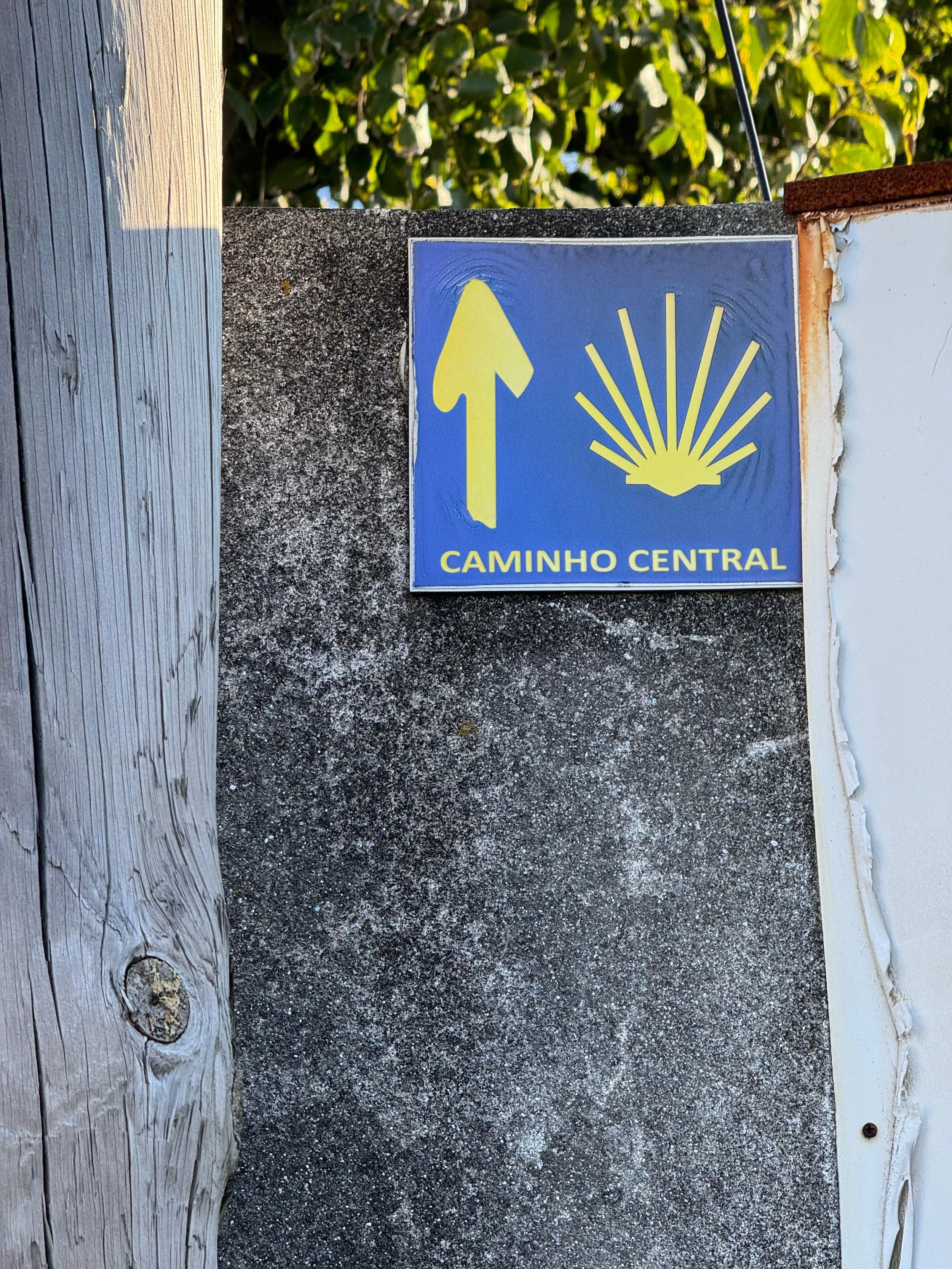 Blue sign with yellow arrow pointing upward and a yellow sunburst symbol, with the words 'CAMINHO CENTRAL' underneath, on a concrete post.
