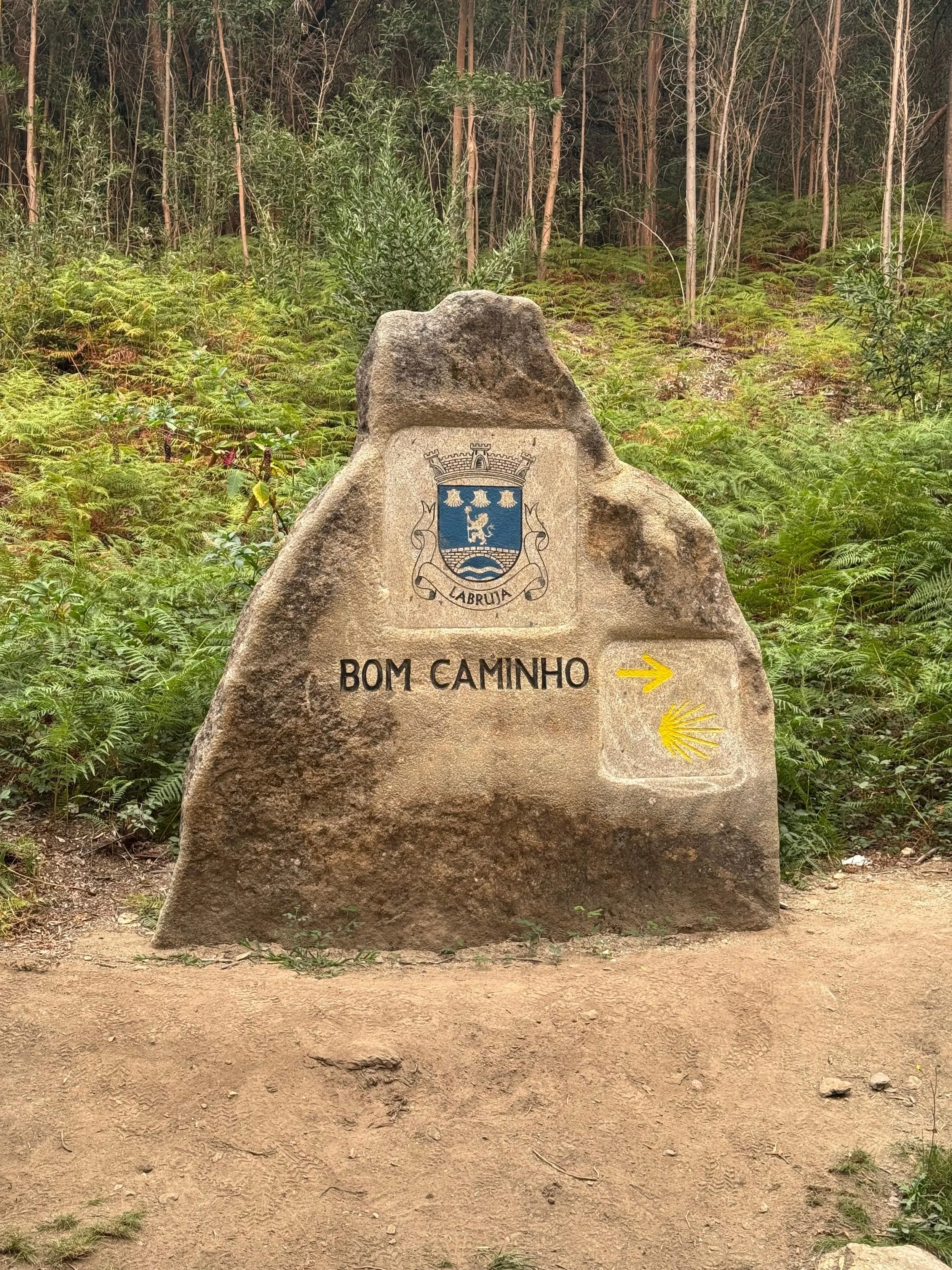 A large rock with a coat of arms, the words 'BOM CAMINHO,' and an arrow pointing to the right with the Shell logo underneath, situated on a dirt trail in a forest.