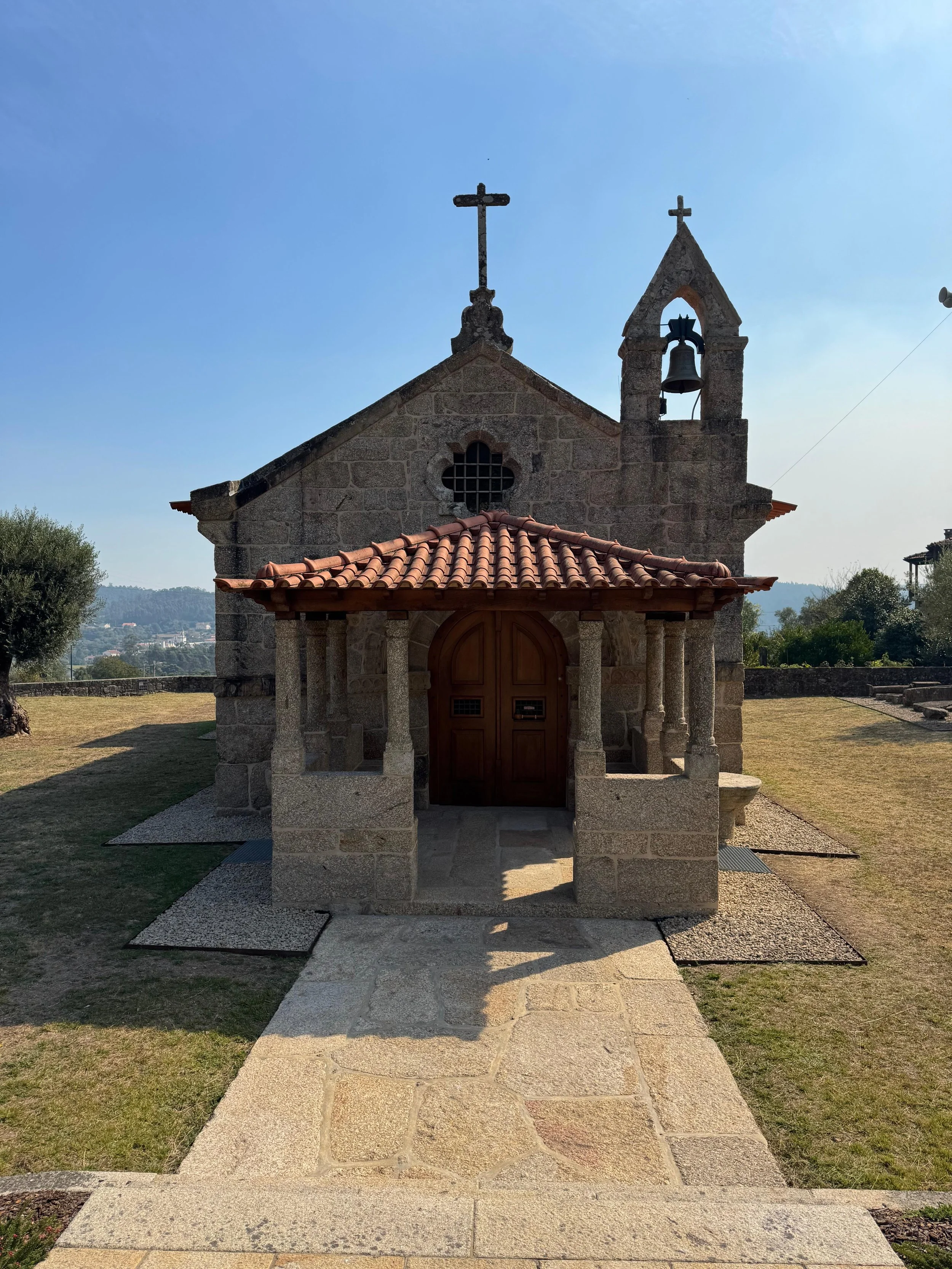 Small stone church with red tile roof and wooden door, surrounded by grass and trees, with crosses on top and a bell tower.