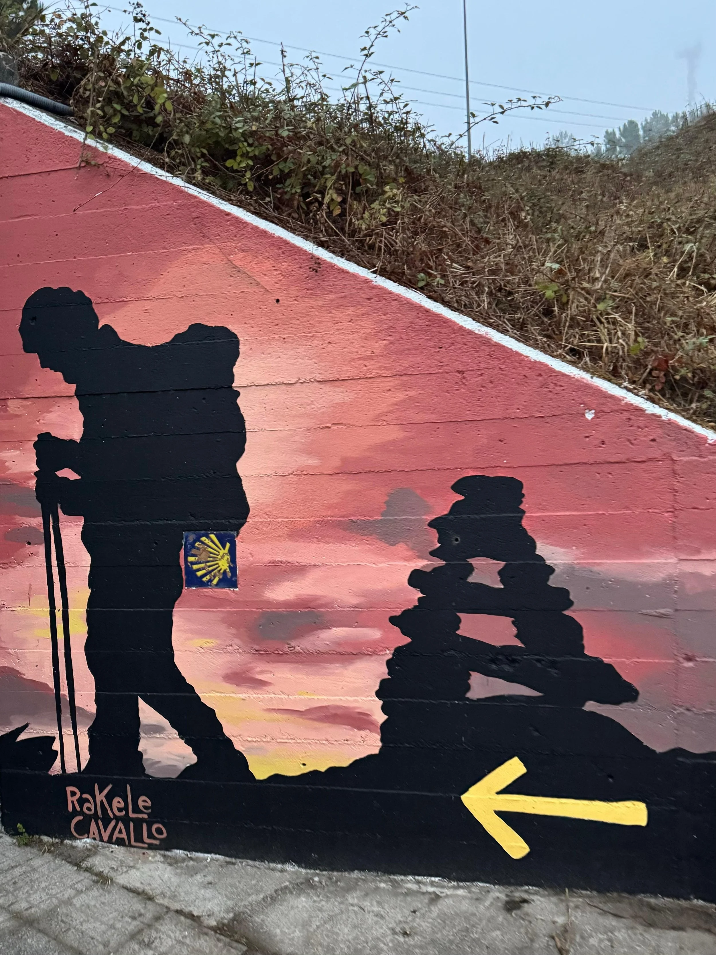 Street mural featuring two hikers with backpacks walking along a trail during sunset, with clouds in the sky. The mural has a pink and orange background, a yellow arrow pointing left, and the artist's signature 'Rakele Cavallo' in the bottom left cor