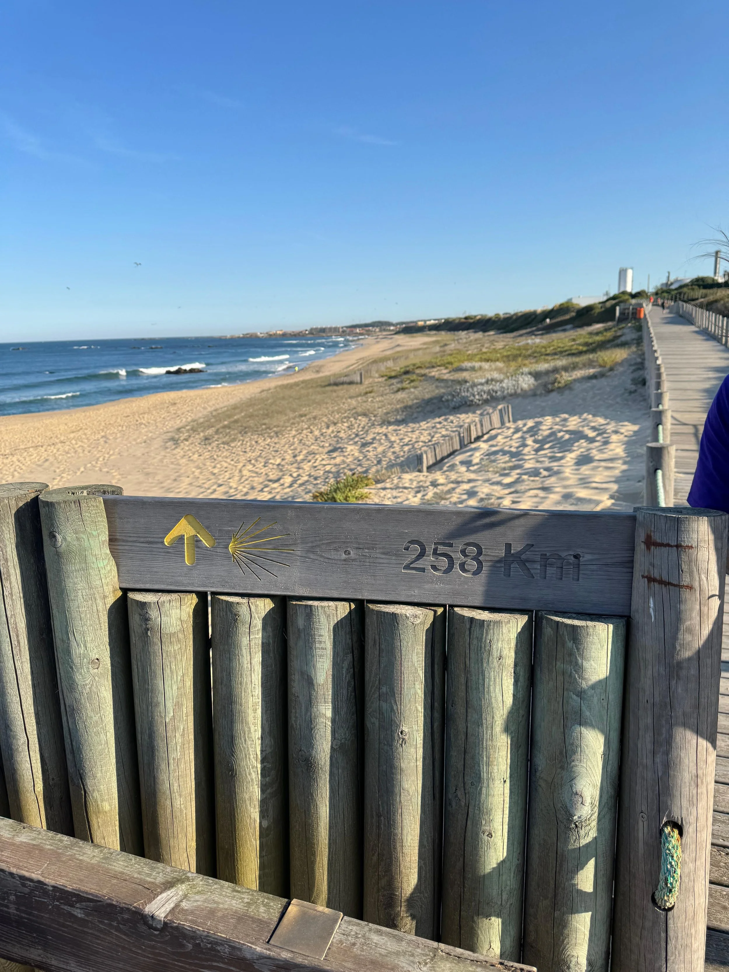 Beach with sandy shore, ocean waves, and a wooden walkway with a sign indicating 258 km to an unspecified location.