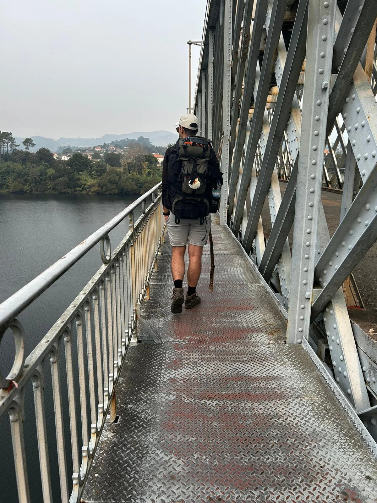 Man walking on a metal bridge over a body of water, carrying a large backpack and wearing hiking gear.