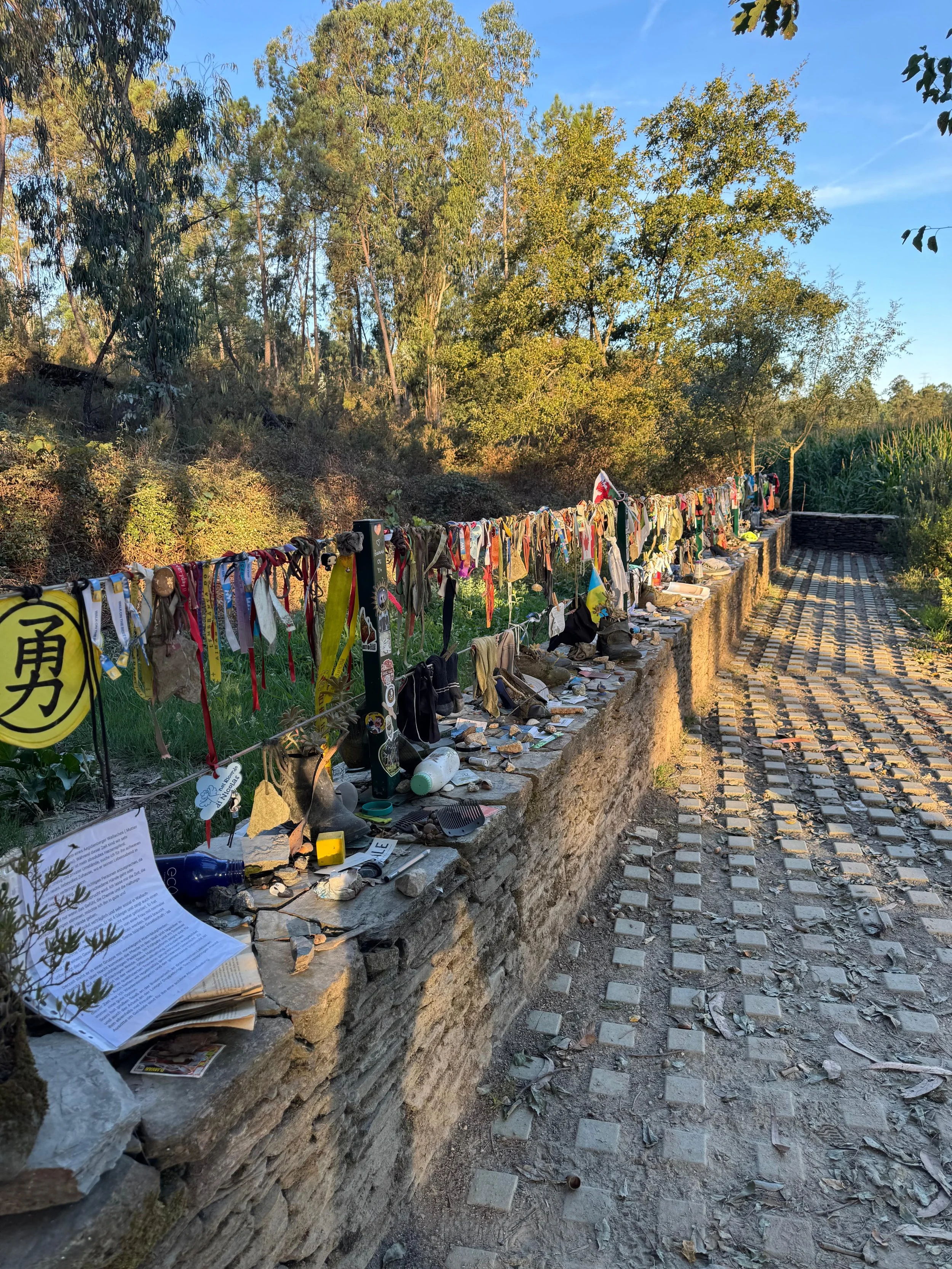 A memorial wall with various items such as ribbons, photographs, and notes placed along a stone ledge, with trees and a blue sky in the background.