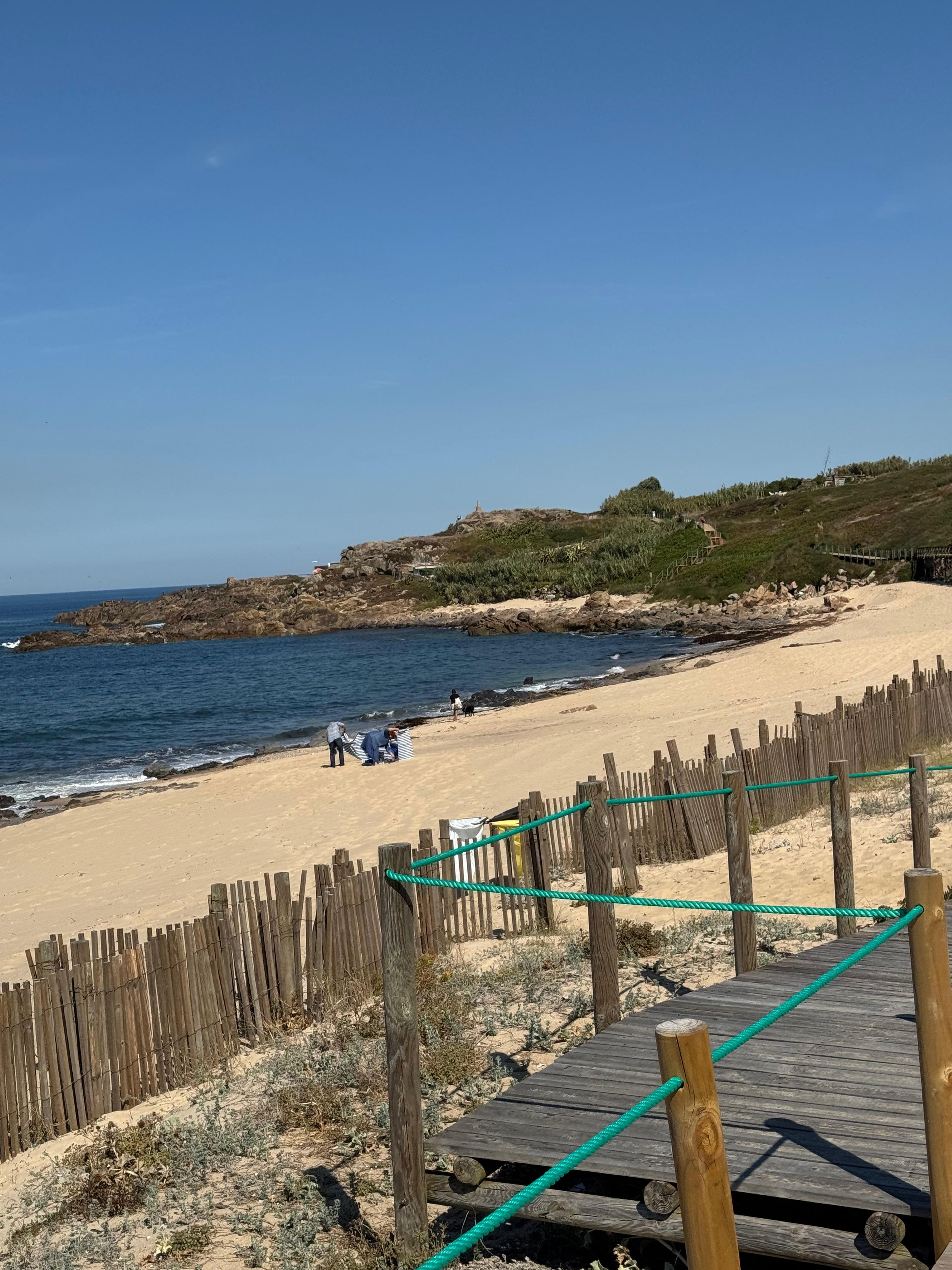 A beach with golden sand, a wooden walkway with green rope railings, a few people near the shoreline, and rocky formations with greenery on hills in the background under a clear blue sky.