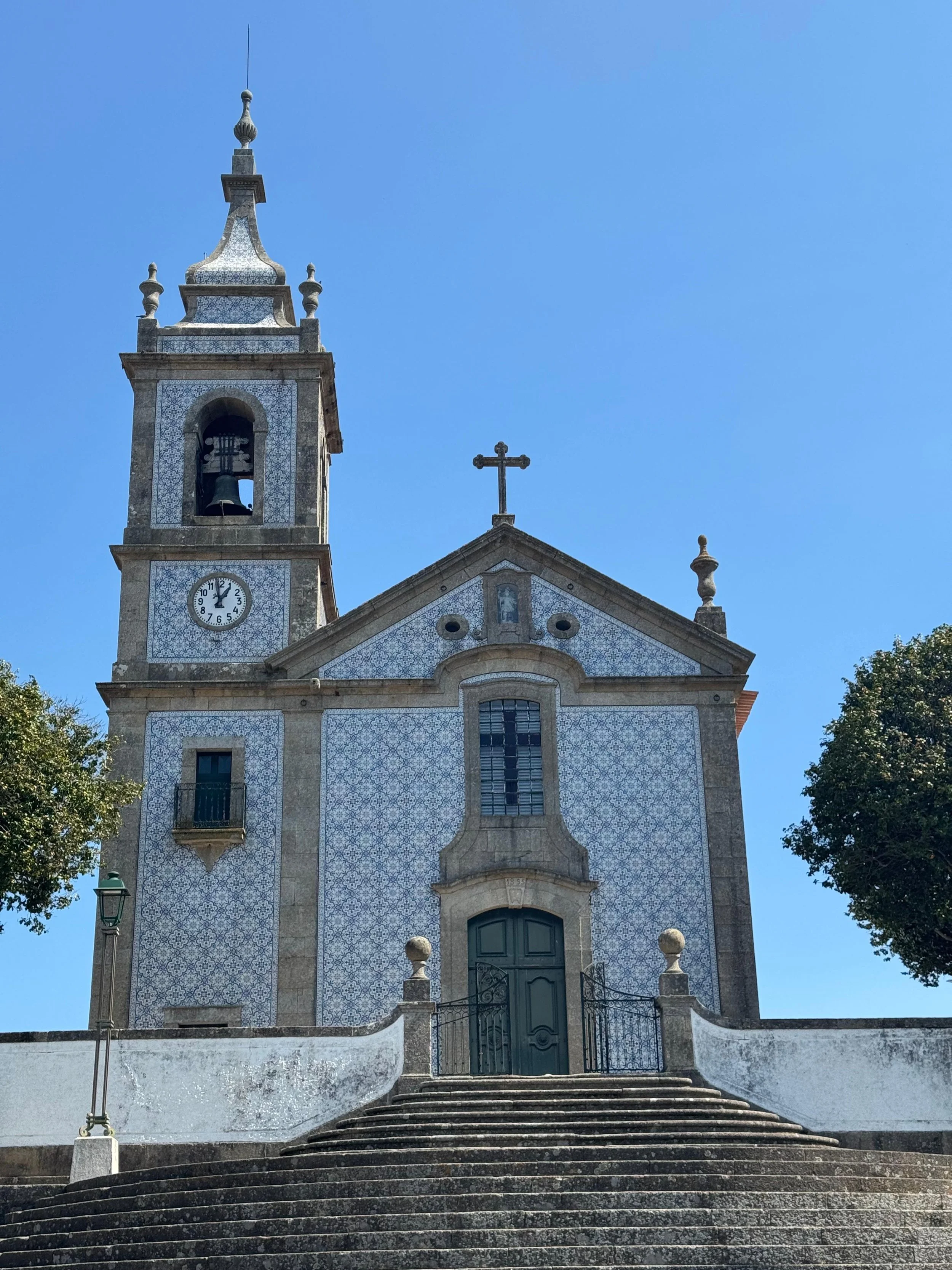 A church building with a bell tower, clock, and cross, accessed by a staircase, under a clear blue sky.