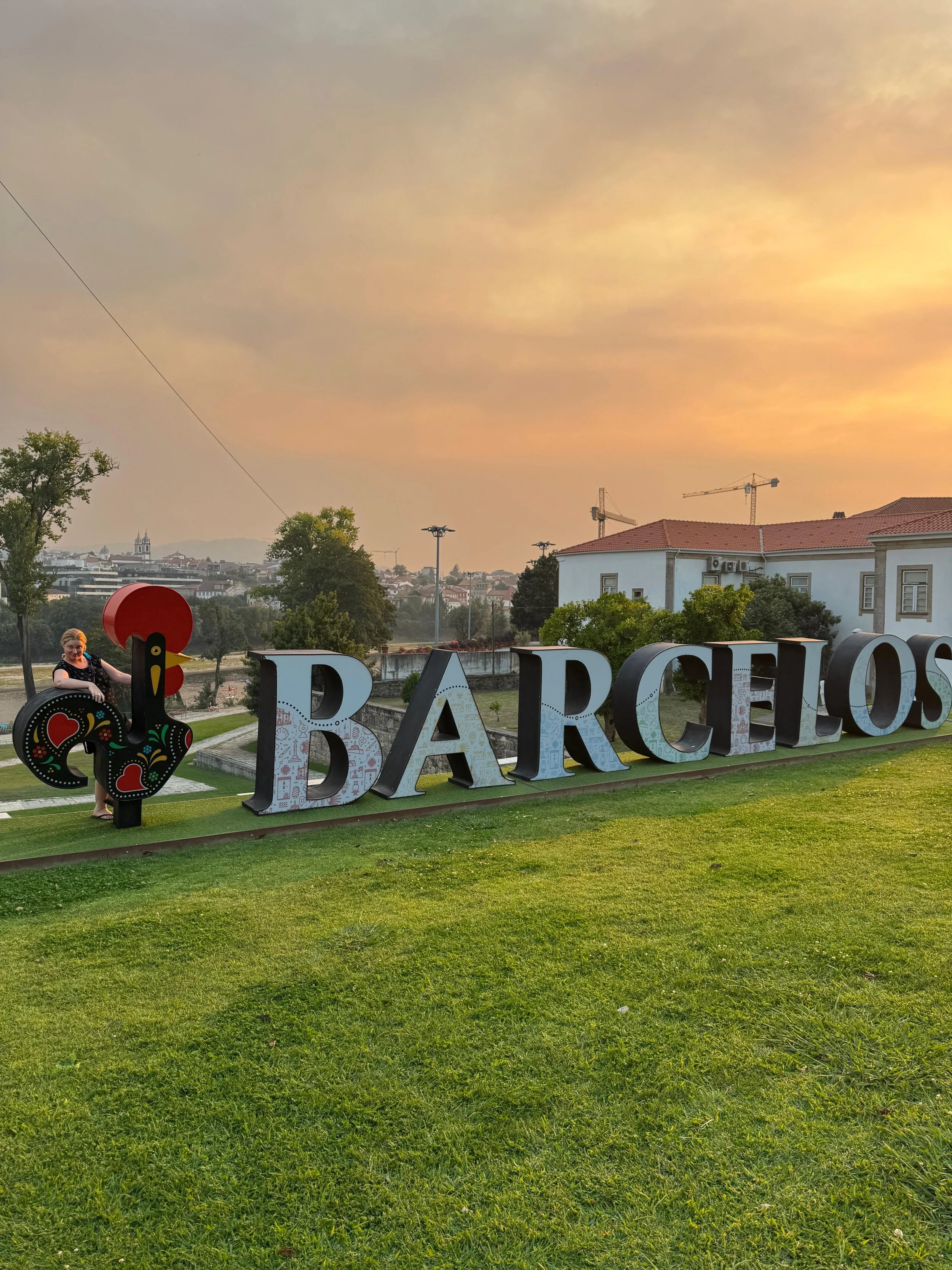 Large sign spelling 'BARCELOS' on a grassy area at sunset with a woman standing next to the letter 'B', some trees, buildings, and construction cranes in the background.