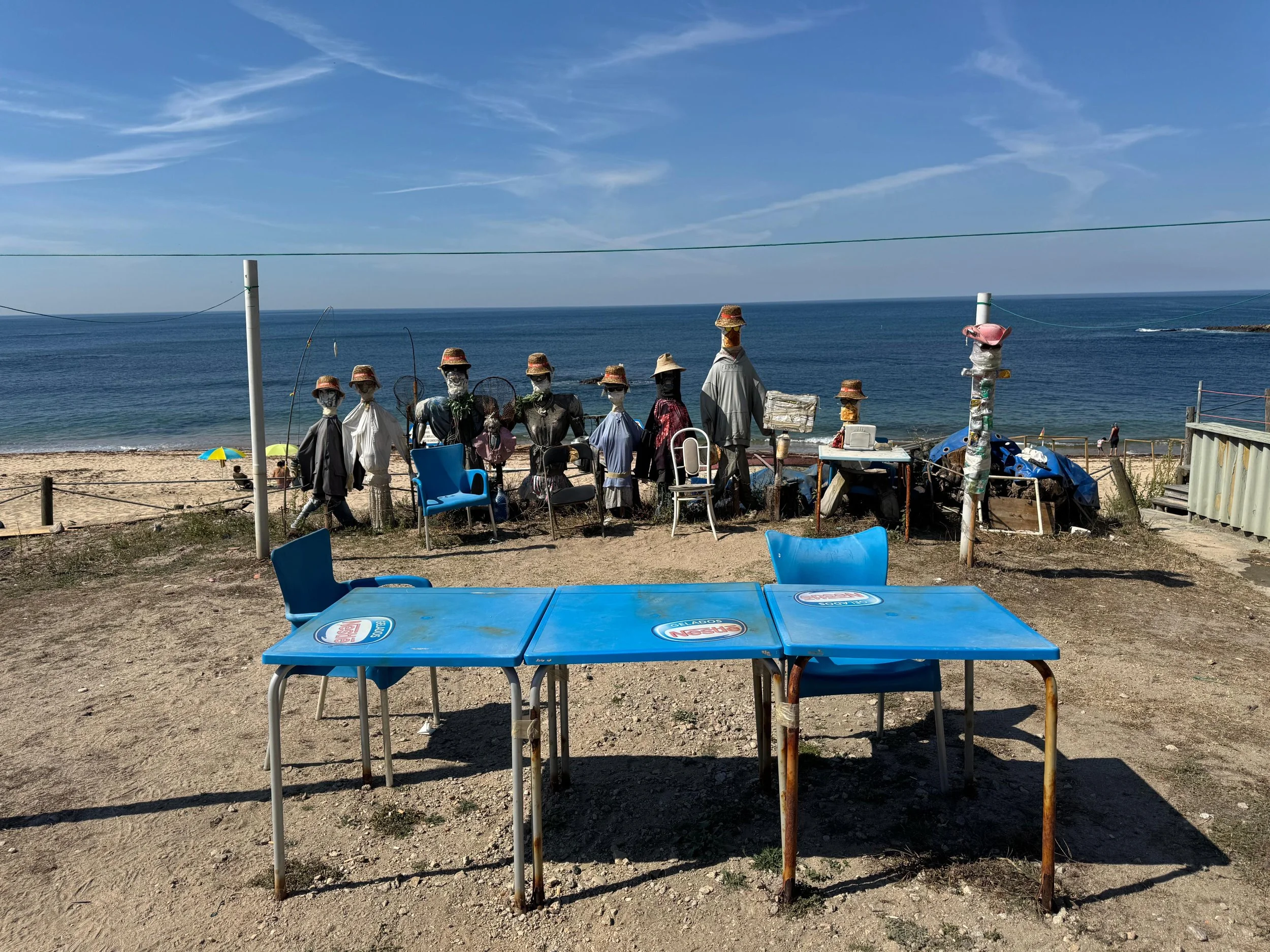 Beachside display of mannequins wearing hats and masks, set up behind blue tables with social distancing chairs in front, near the ocean with sandy shores and blue sky.