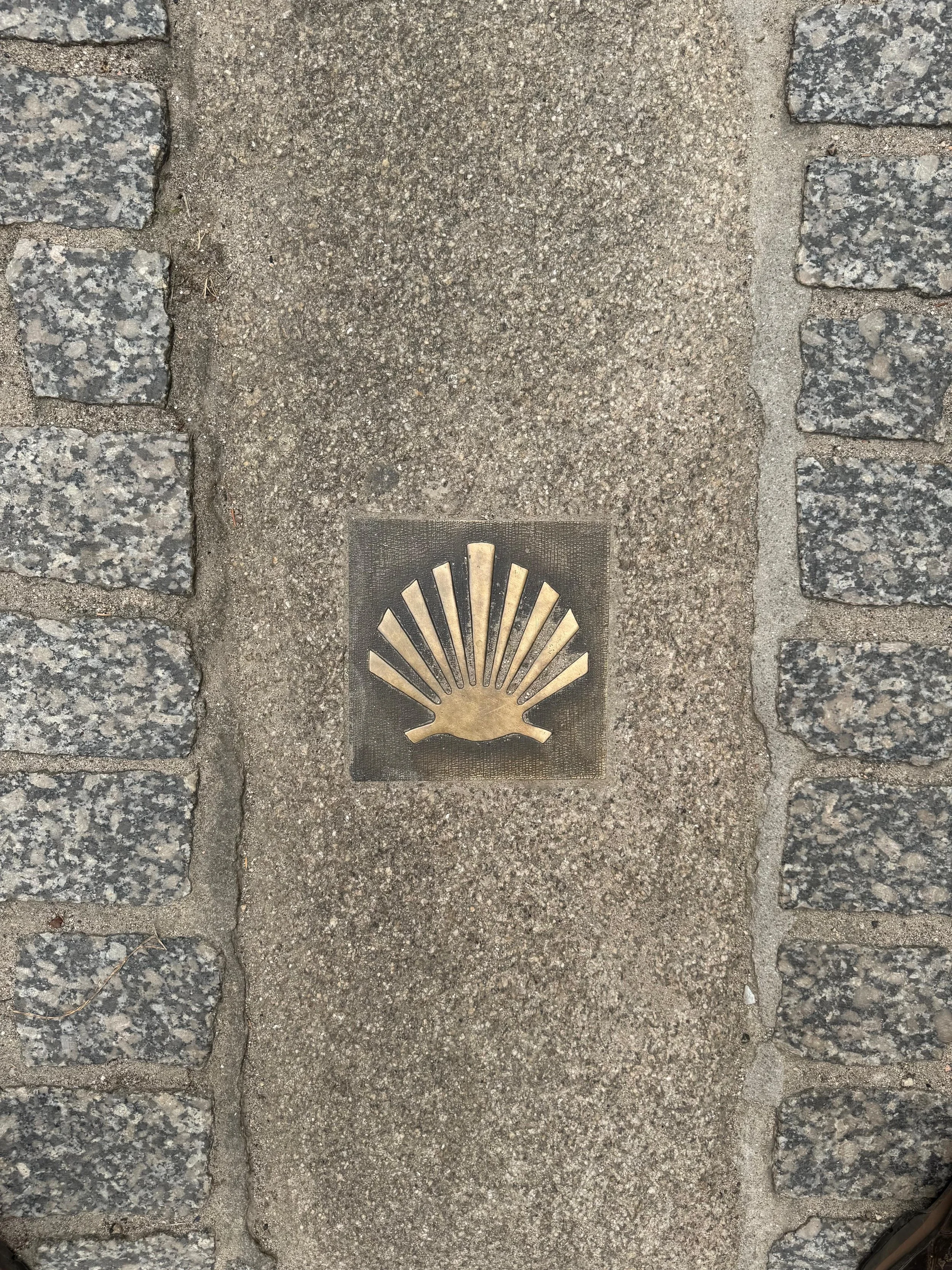 A bronze inlaid emblem of a scallop shell on the pavement, flanked by two pebbled stone borders.
