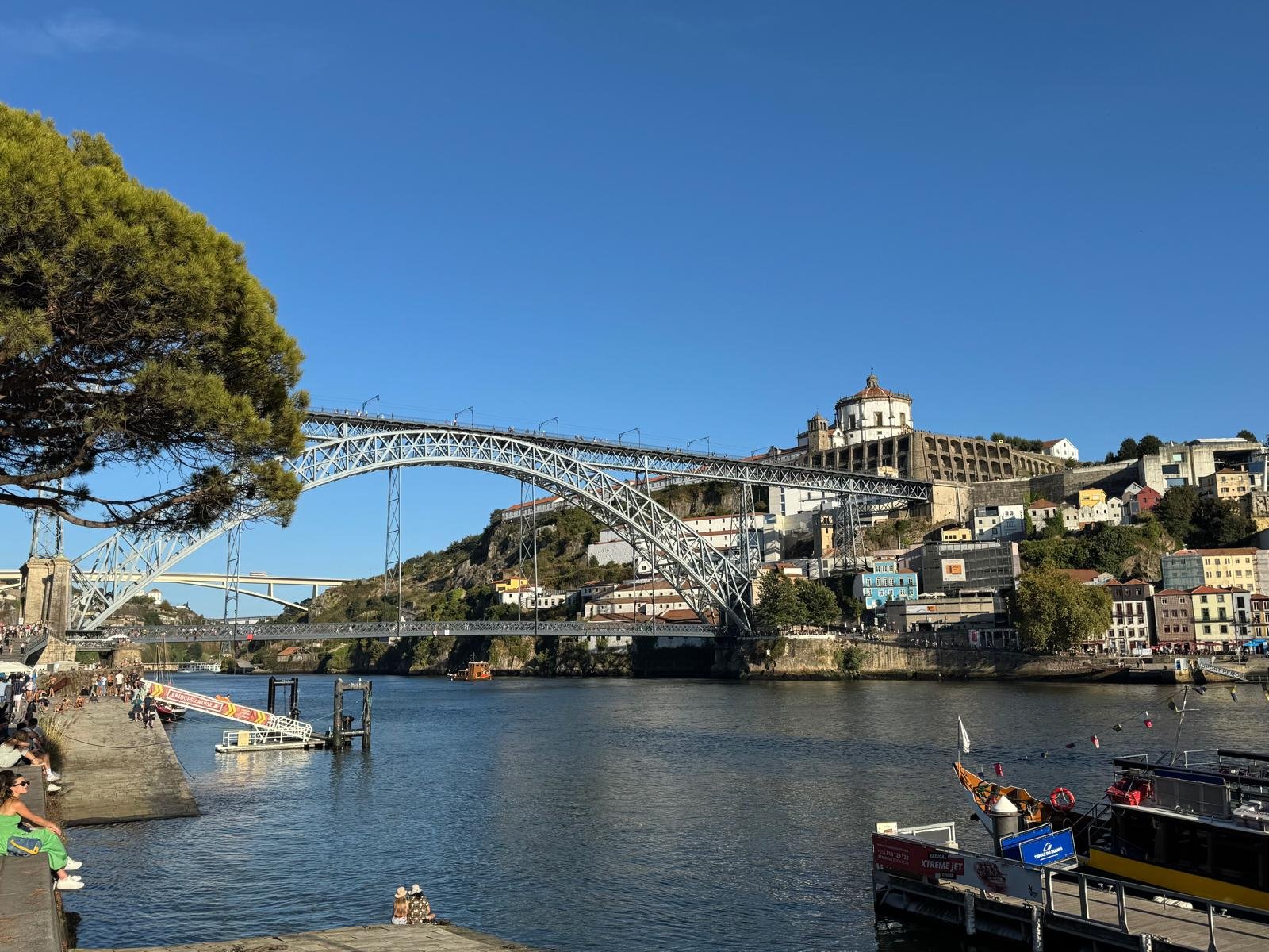 Ein malerischer Blick auf eine Stadtlandschaft mit einer großen Bogenbrücke, die einen Fluss überspannt, bunten Gebäuden, die sich einen Hang hinaufziehen, und einem strahlend blauen Himmel.