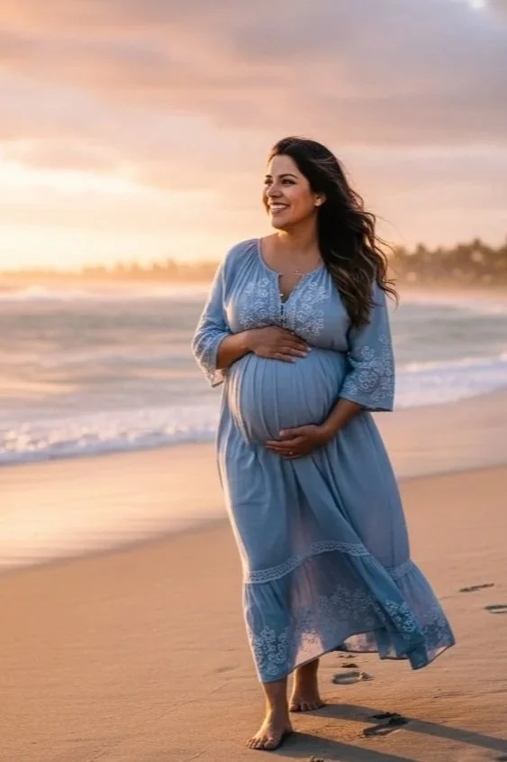 Pregnant woman in a light blue dress walking barefoot on the beach during sunset, smiling and holding her belly.