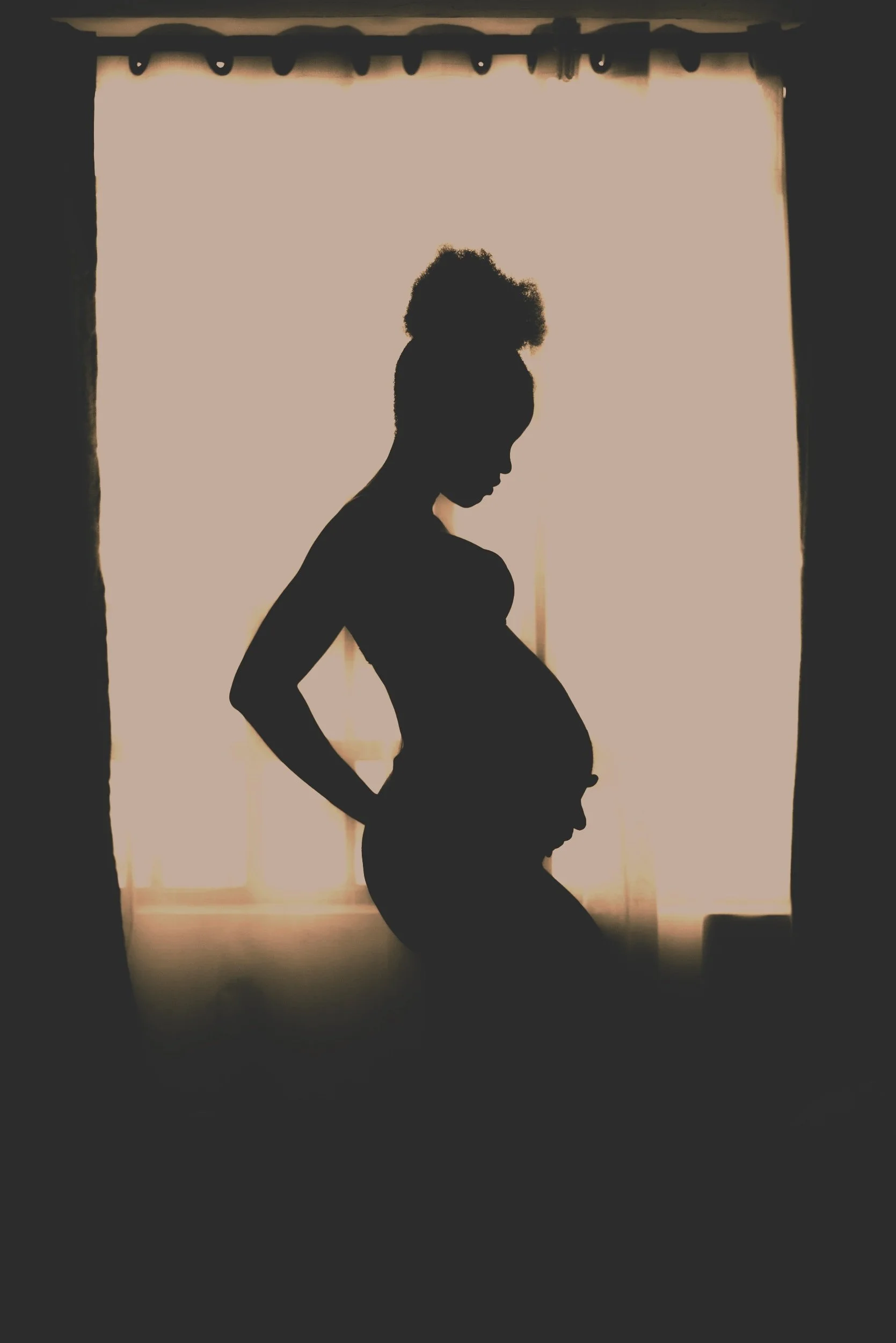 Silhouette of a pregnant woman with her hand on her back, standing in front of a window with curtains.