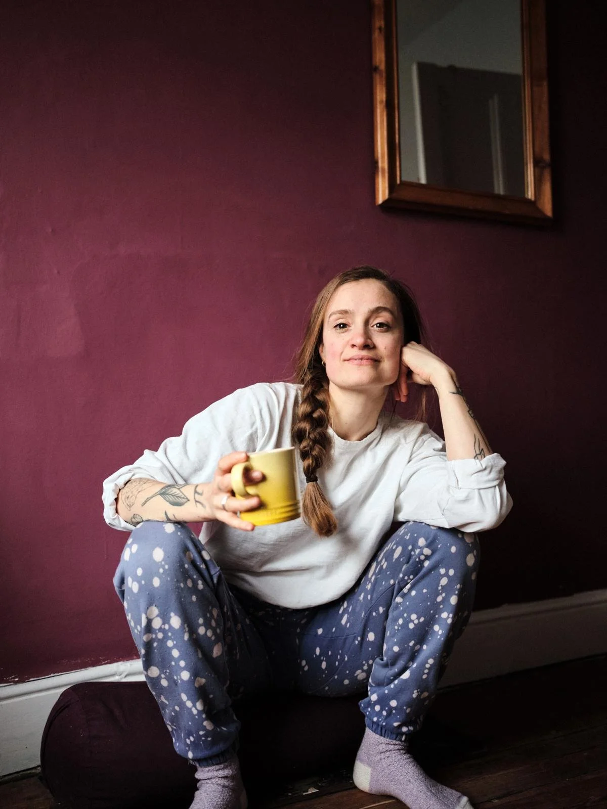 A woman with tattoos on her arms and a braid, sitting cross-legged on a cushion on the floor, holding a yellow mug, with a relaxed expression against a purple wall with a mirror.