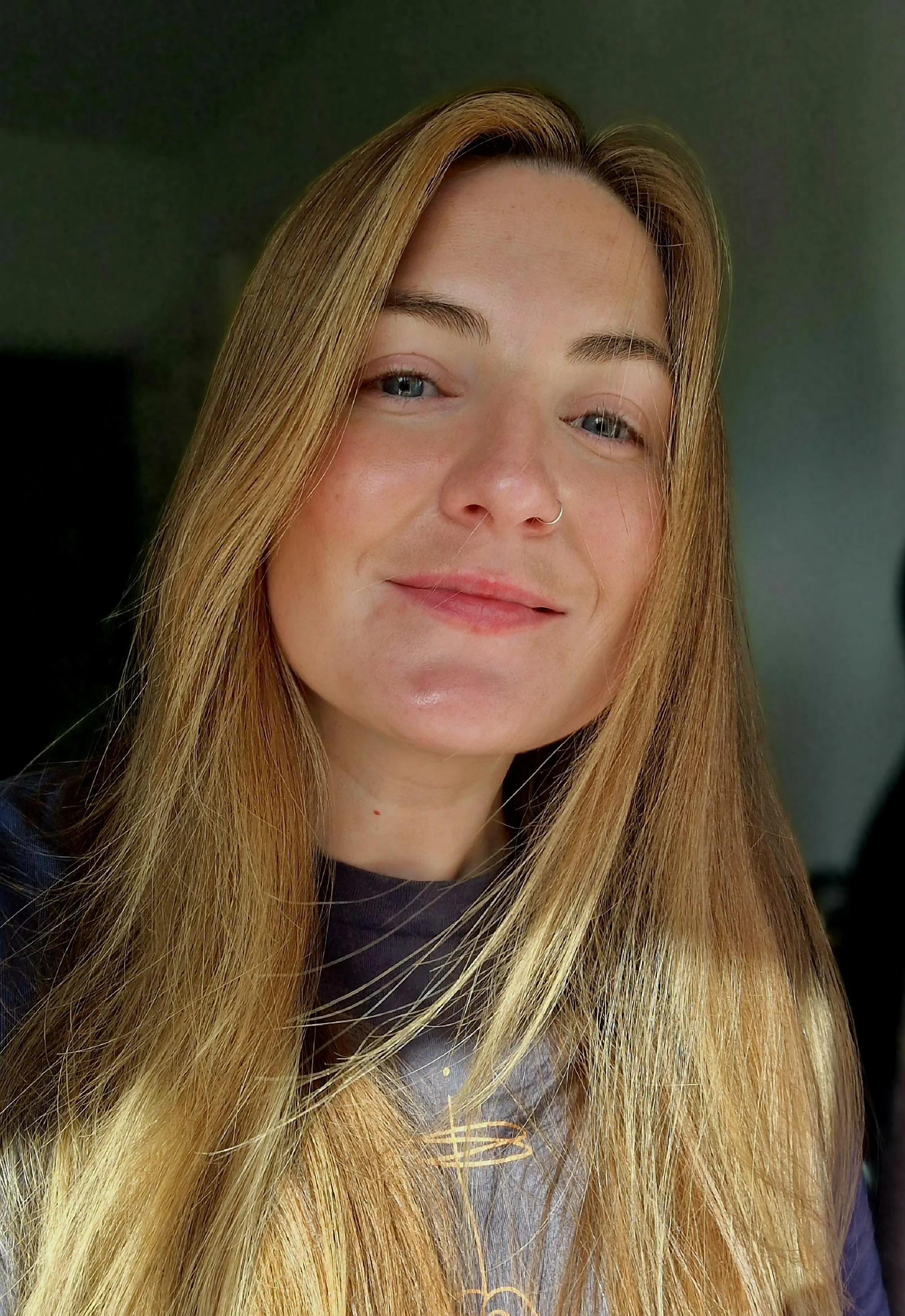 Close-up of a woman with long blond hair, blue eyes, and a nose ring, smiling gently.