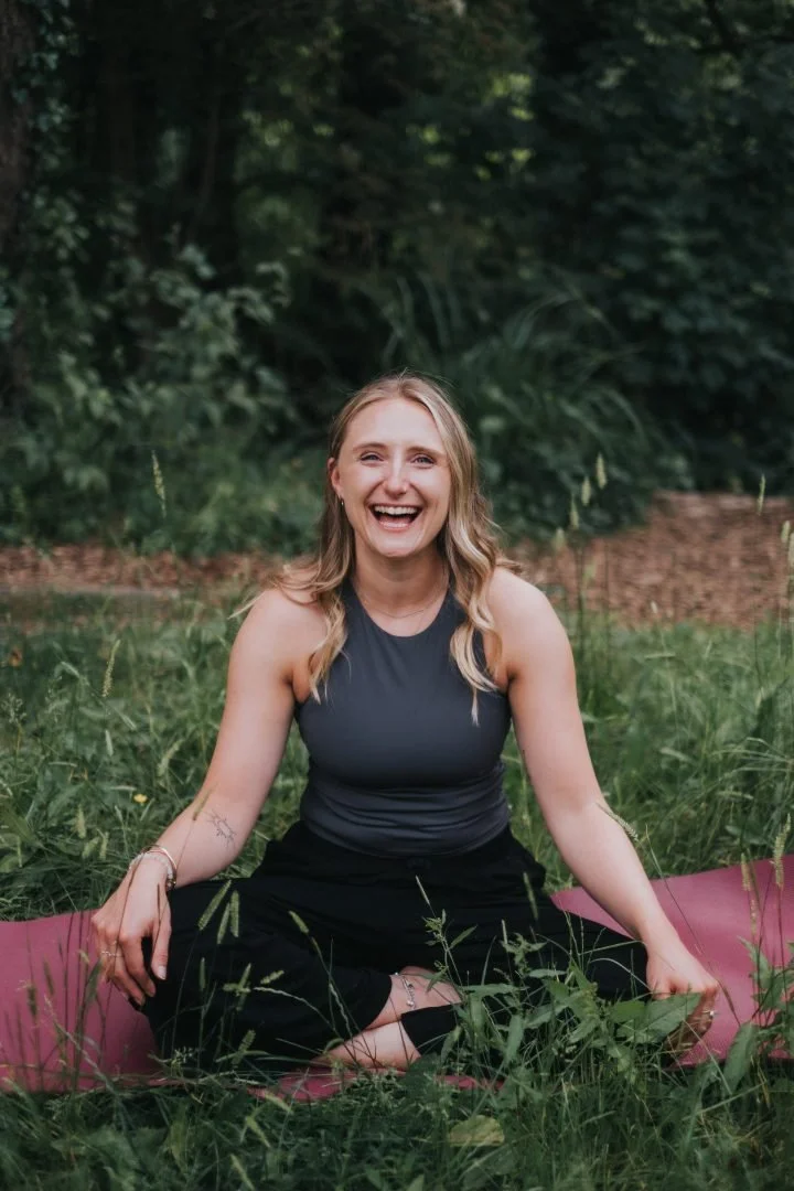 A woman sitting on a pink yoga mat outdoors, smiling widely, surrounded by green grass and trees in the background.