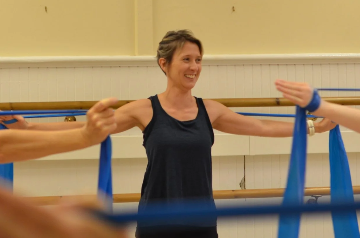 Woman in a black tank top smiling while participating in a fitness class, stretching with resistance bands at a gym or dance studio.