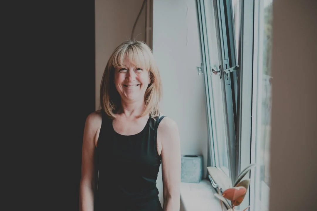 A smiling woman with shoulder-length blonde hair wearing a black sleeveless top standing near an open window with daylight streaming in.