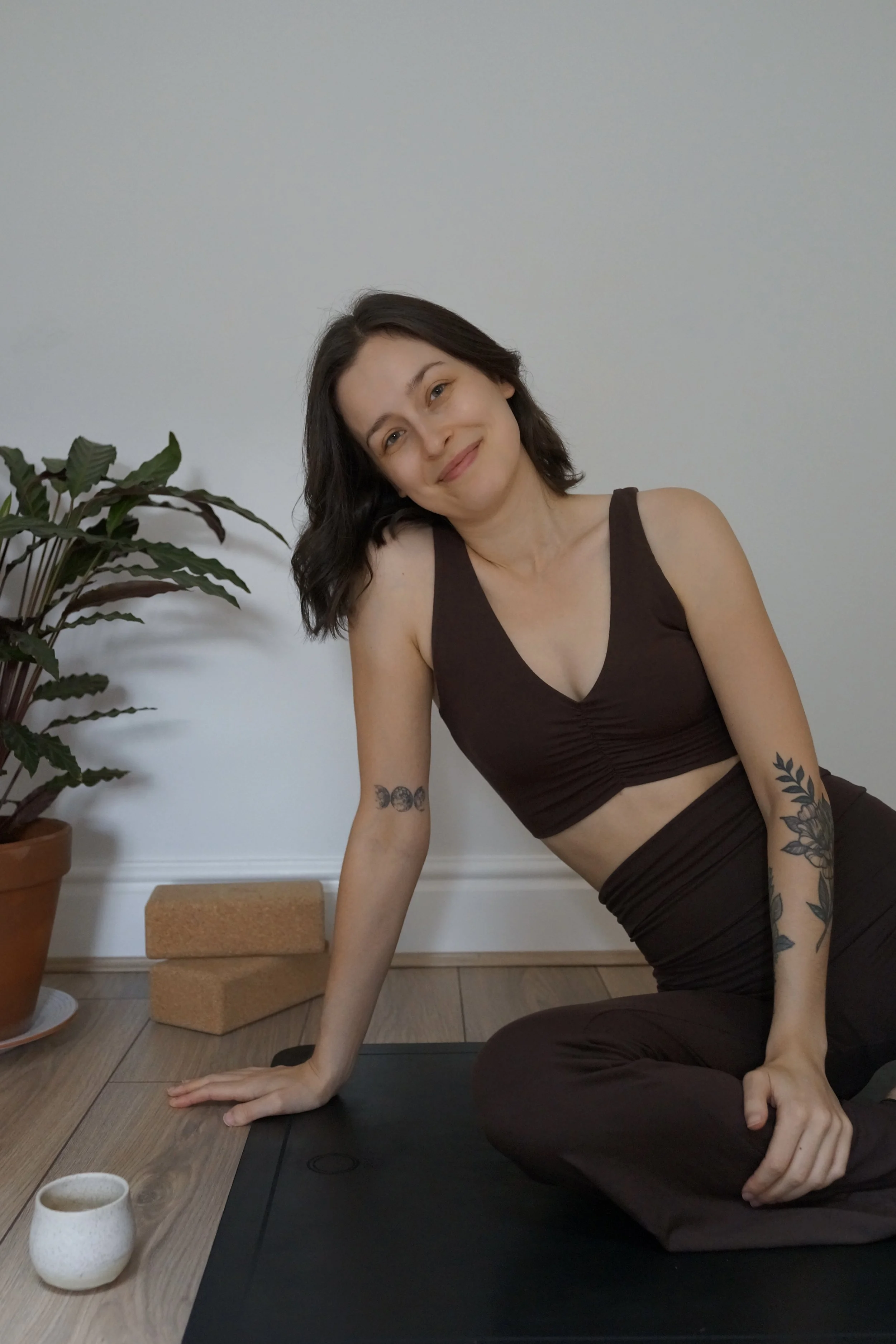 A woman with dark hair sitting on a yoga mat, smiling, in a room with wooden flooring and a white wall, near a potted plant and a cup of tea.