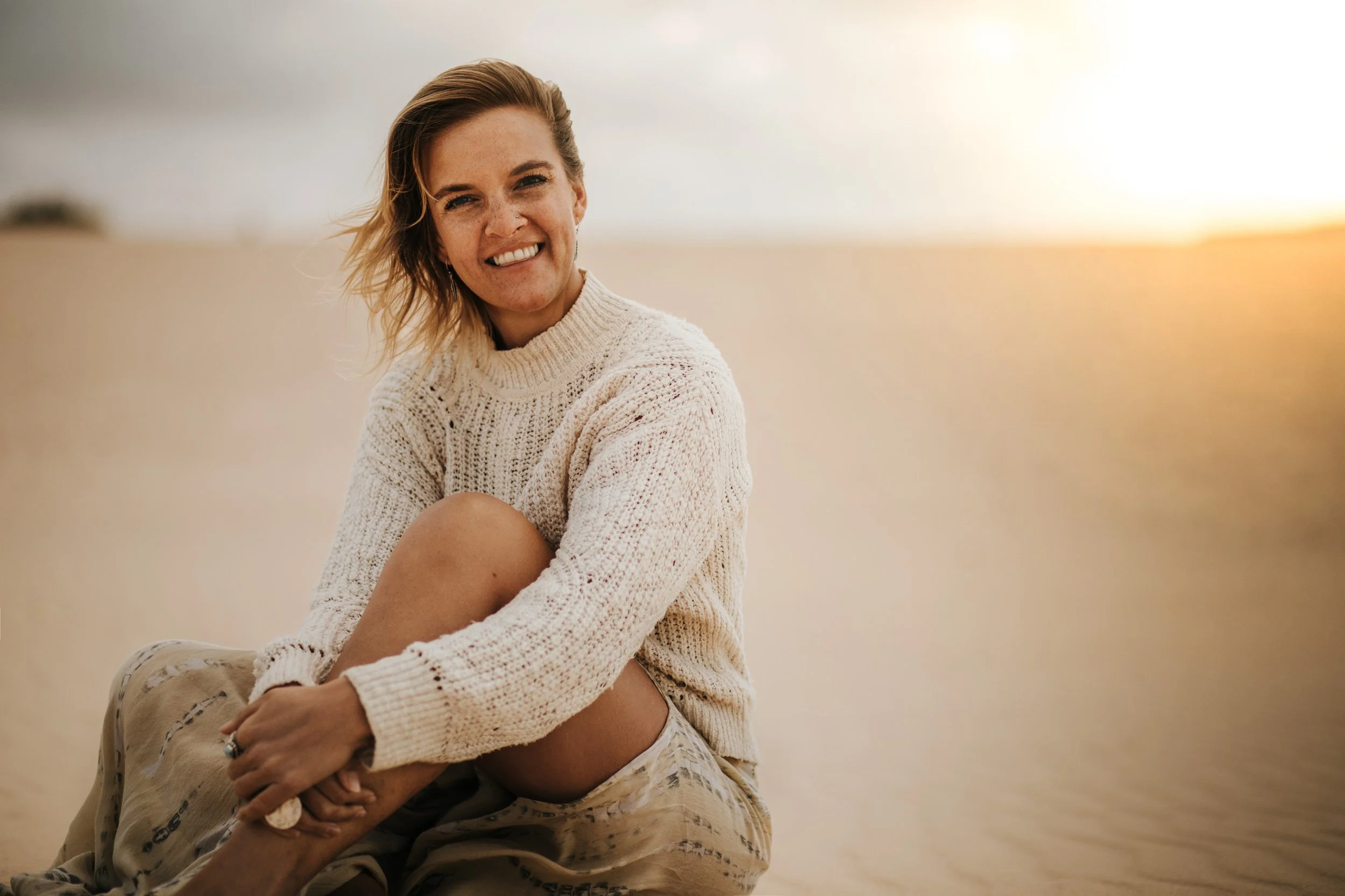 A woman with short blonde hair sitting on a sandy beach at sunset, smiling and wearing a cream sweater and patterned pants.