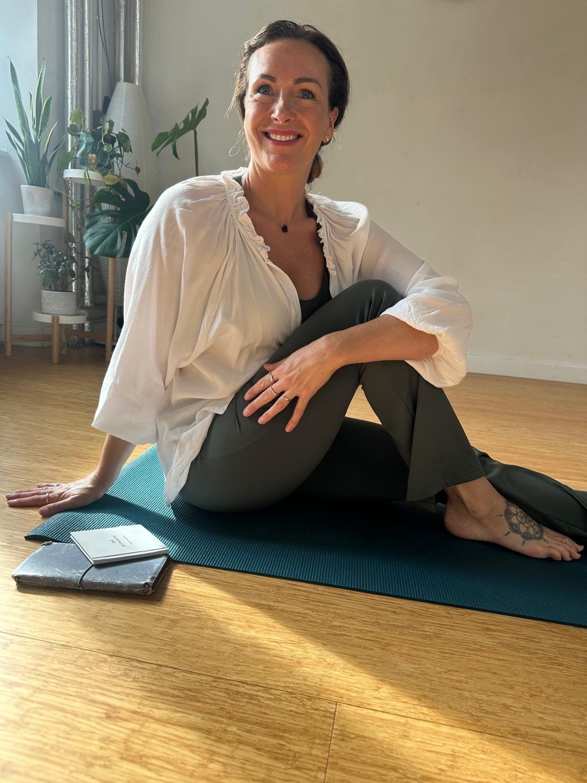 A woman sitting on a yoga mat in a well-lit room, smiling, with potted plants and sunlight in the background.
