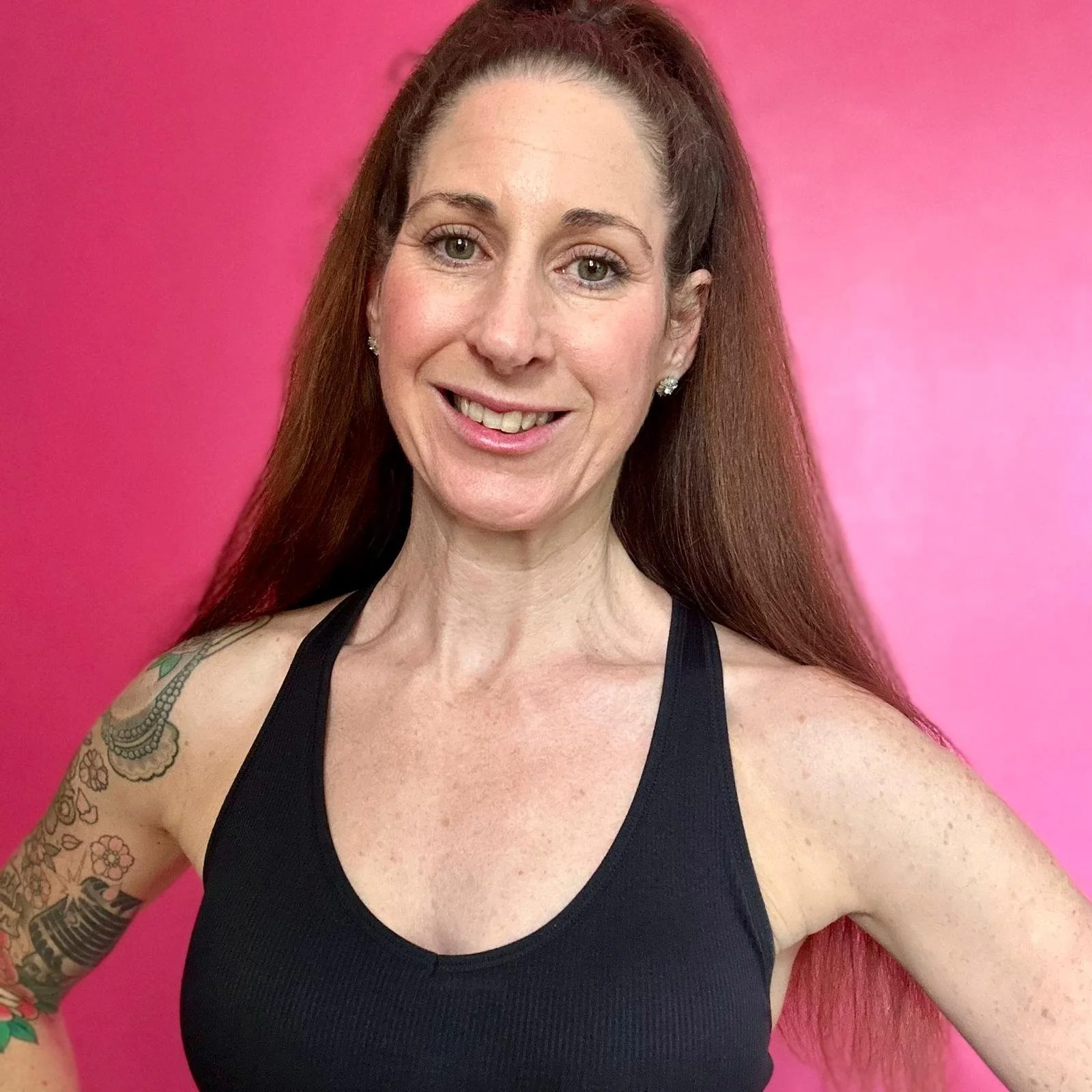A woman with long red hair, wearing a black tank top, smiling, standing against a pink background.