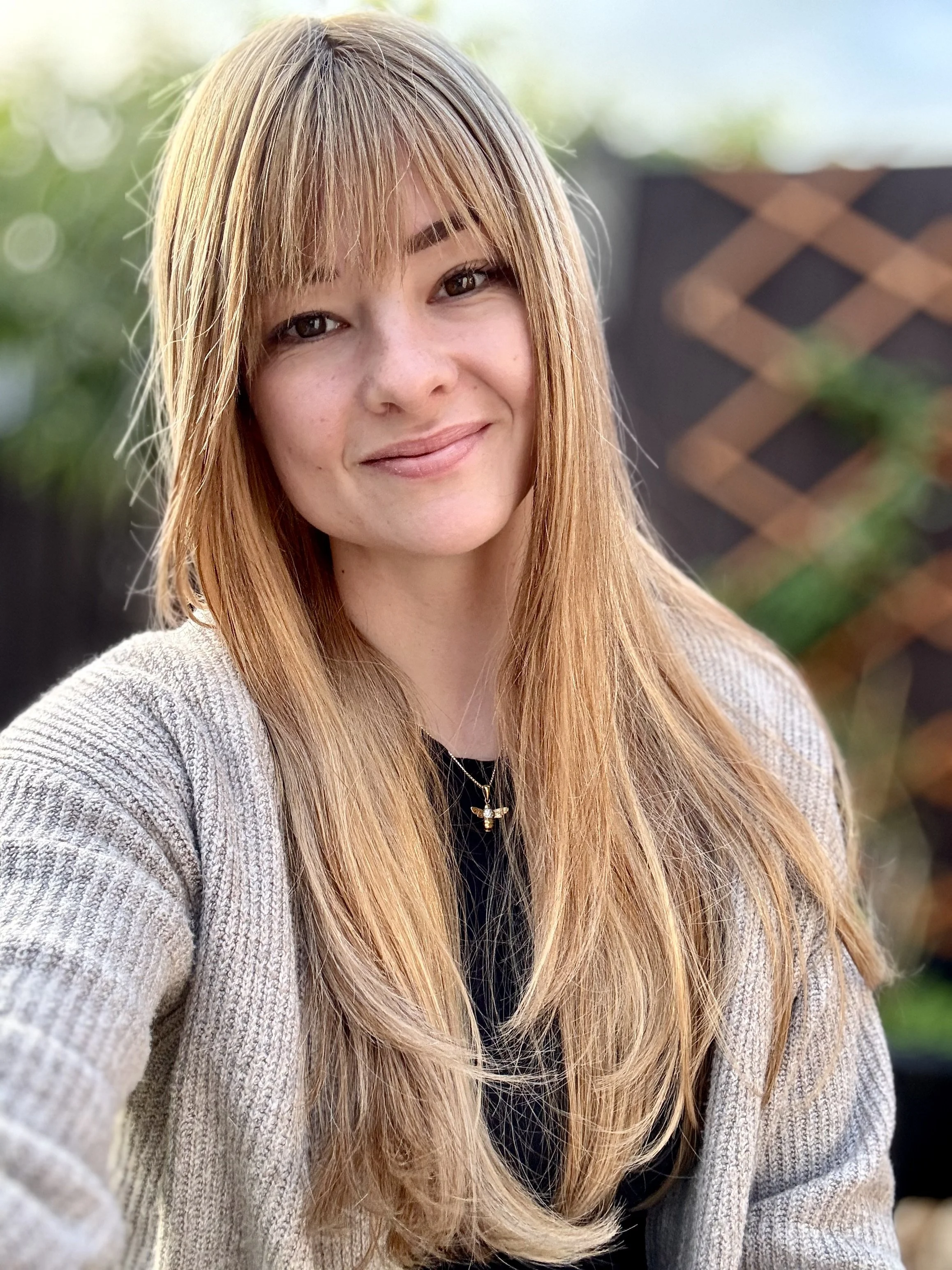 Fee, a female counsellor in her thirties with long blonde hair and a fringe smiles gently and welcoming into the camera. The background is blurred but you can just see greenery like trees and climbing plants.