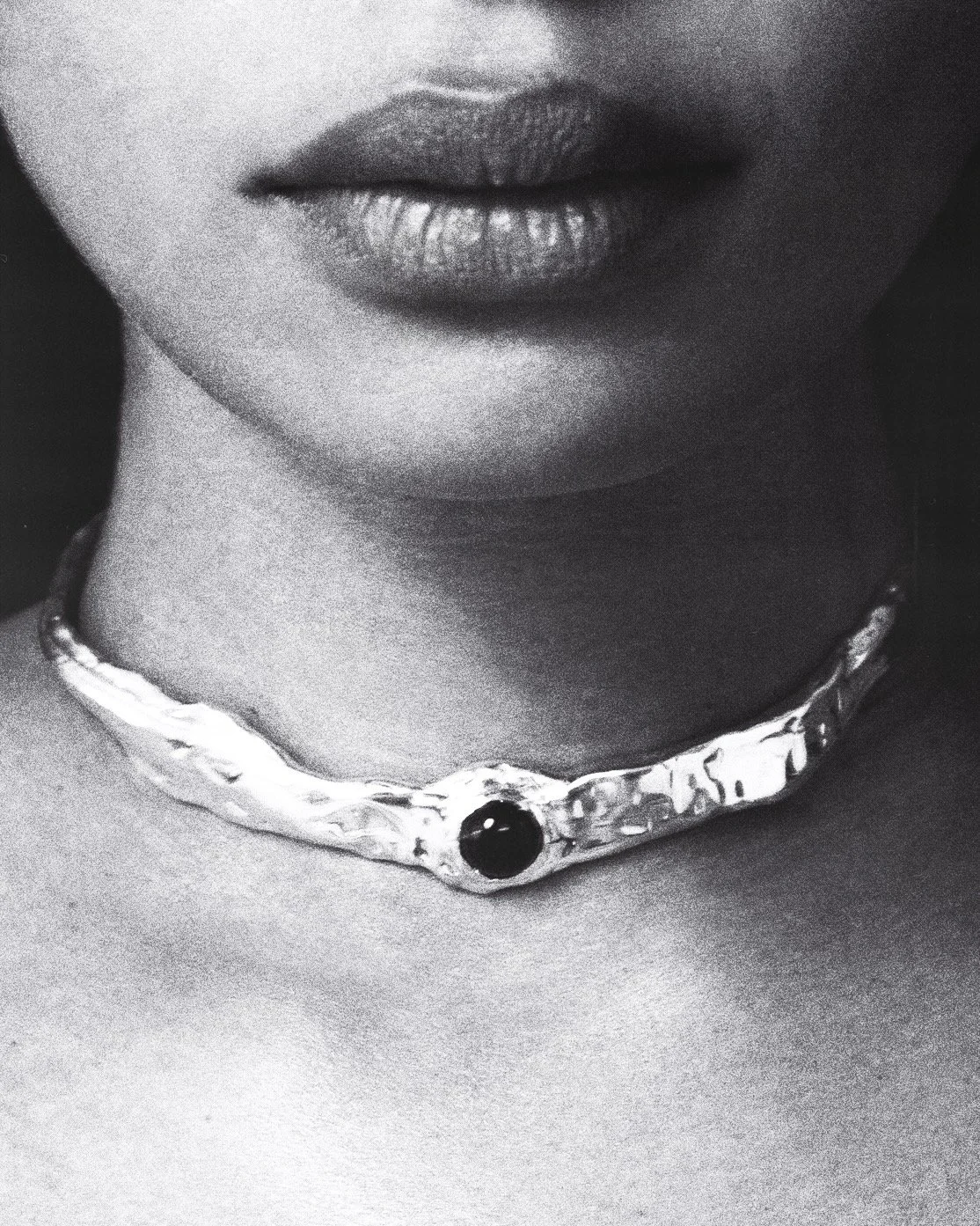 Close-up of a person's lips and neck, decorated with a textured metallic choker with a central dark gemstone, in black and white.