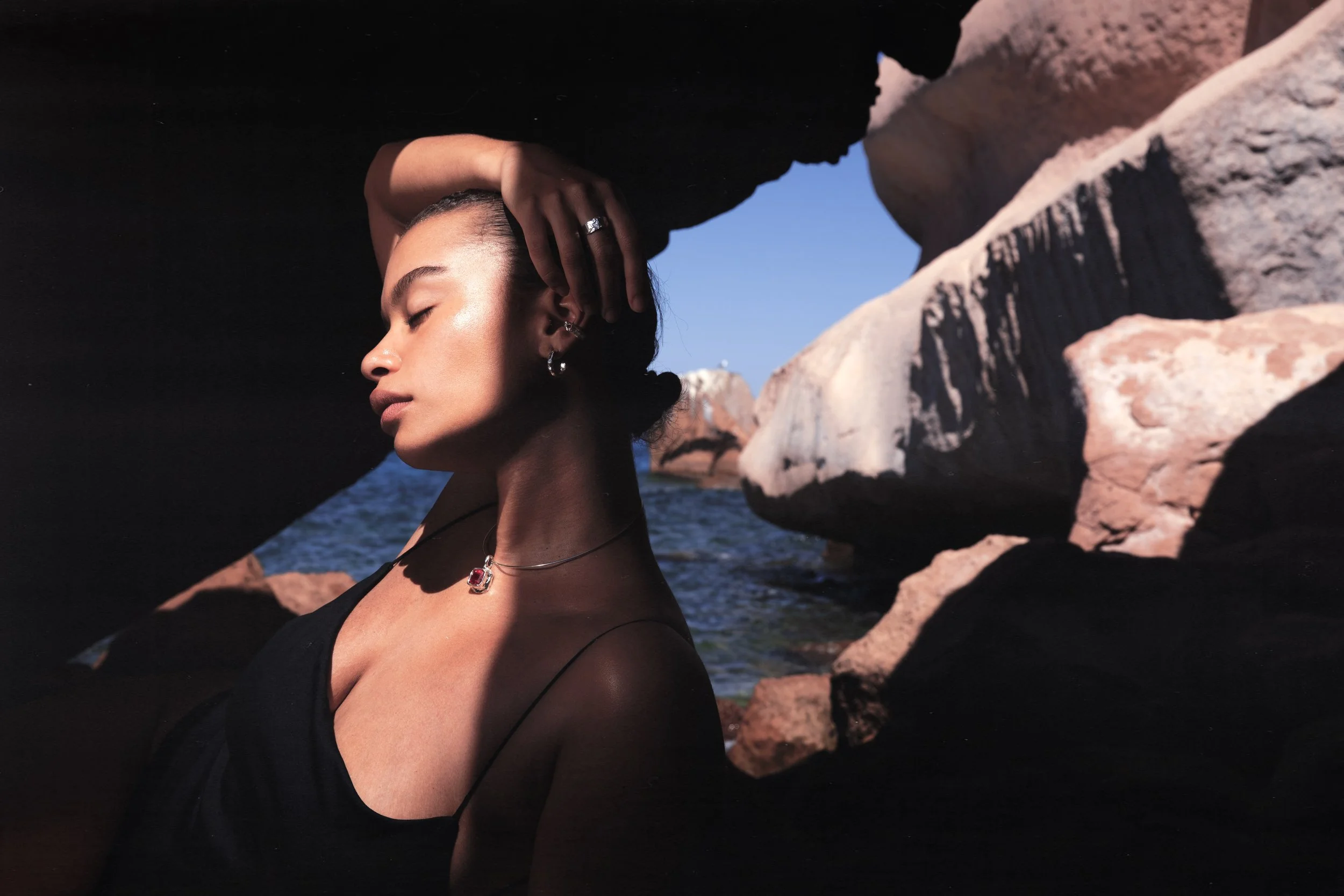 A woman with jewelry and dark clothing stands between large rocks on a coastline, with her eyes closed and her hand resting on her head, under a bright sky.
